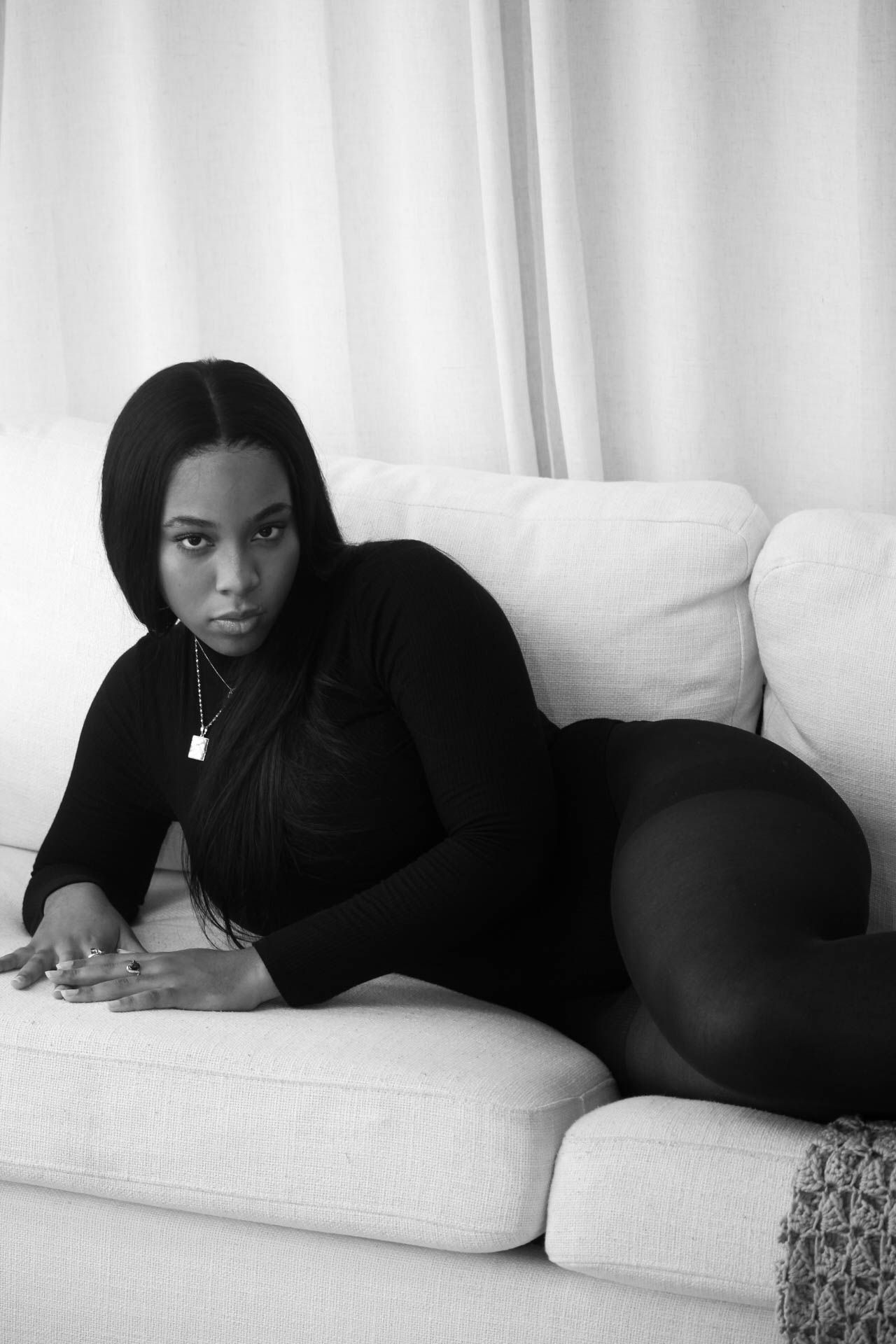Black and white photo of a woman in a black outfit posing on a white sofa with curtains in the background.