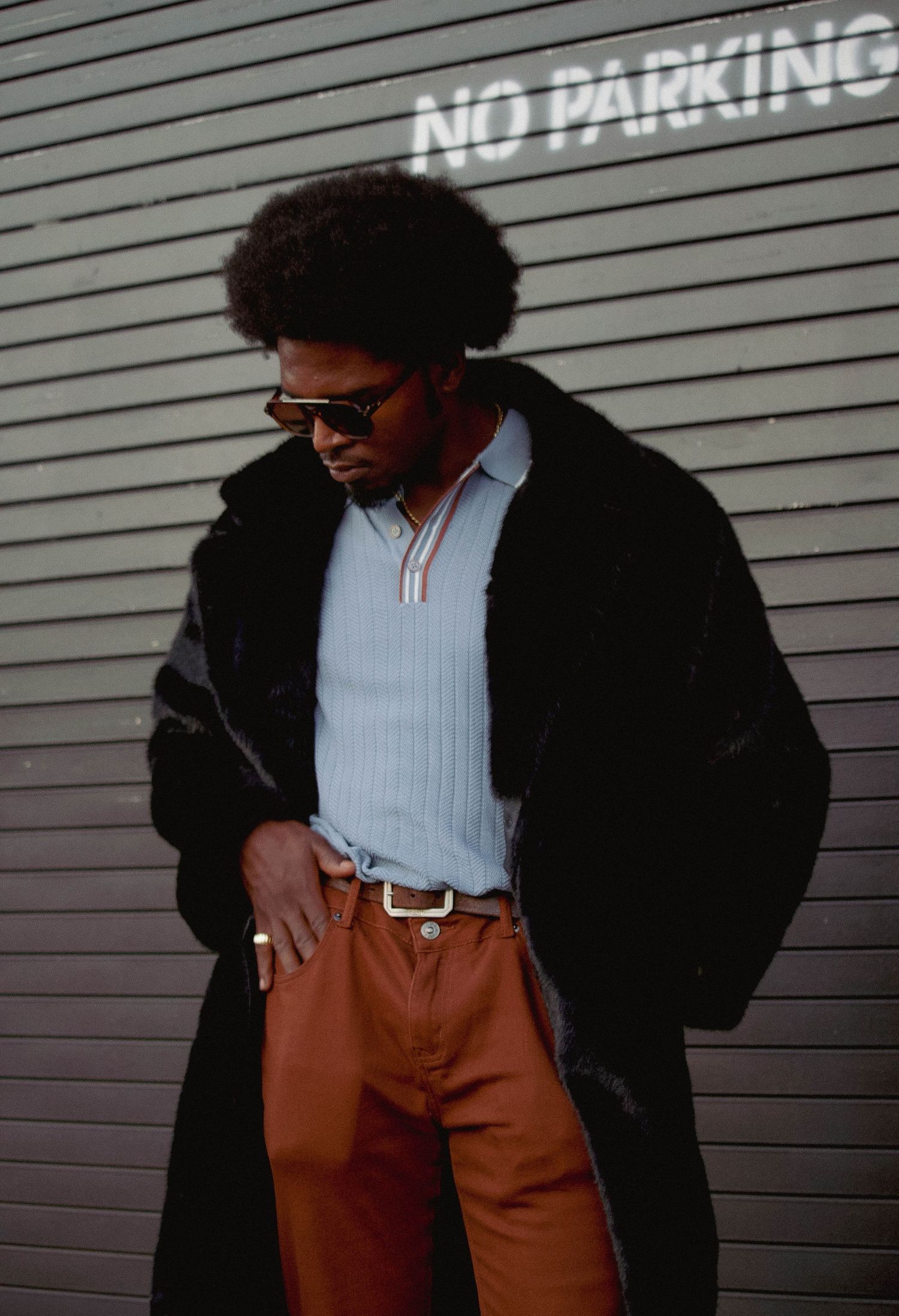 Stylish man with afro wearing black fur coat, blue sweater, brown pants and sunglasses near No Parking sign.
