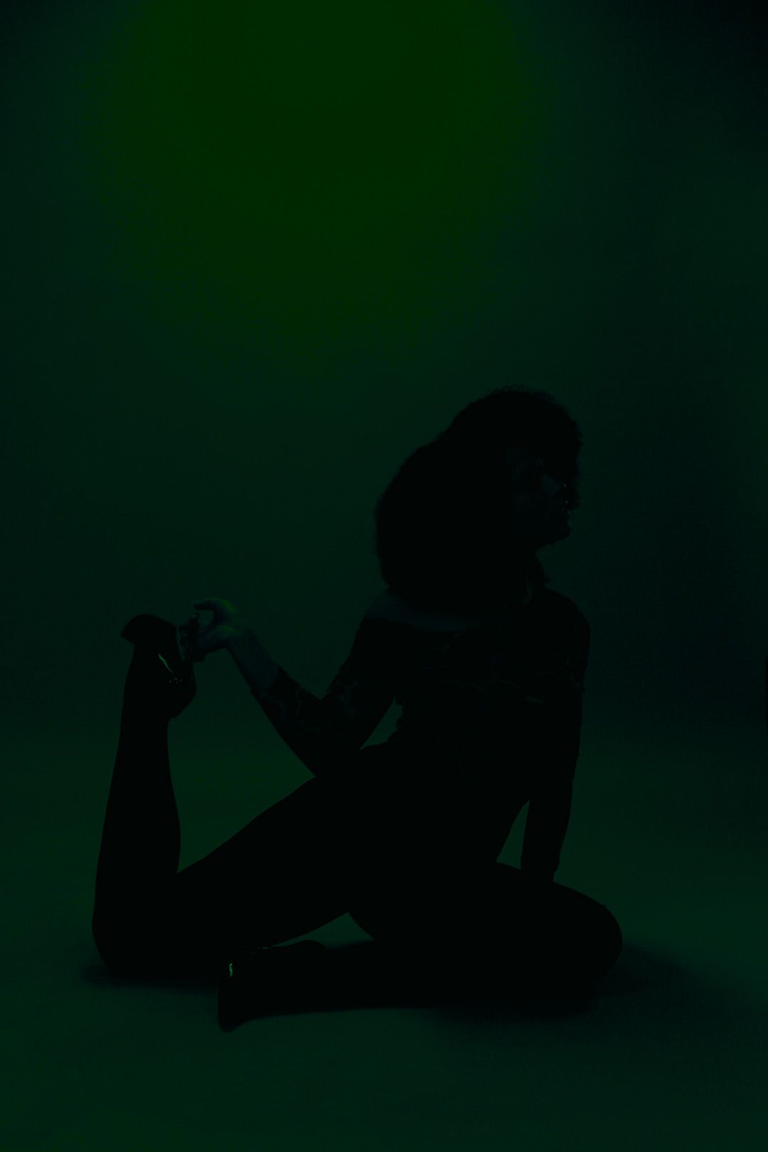 Silhouette of woman sitting on floor holding ankle against dark green background in artistic pose.