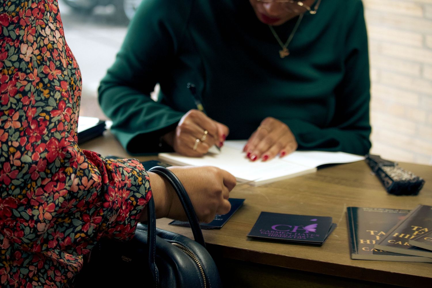 Two people signing documents at a wooden table with books and a phone nearby.