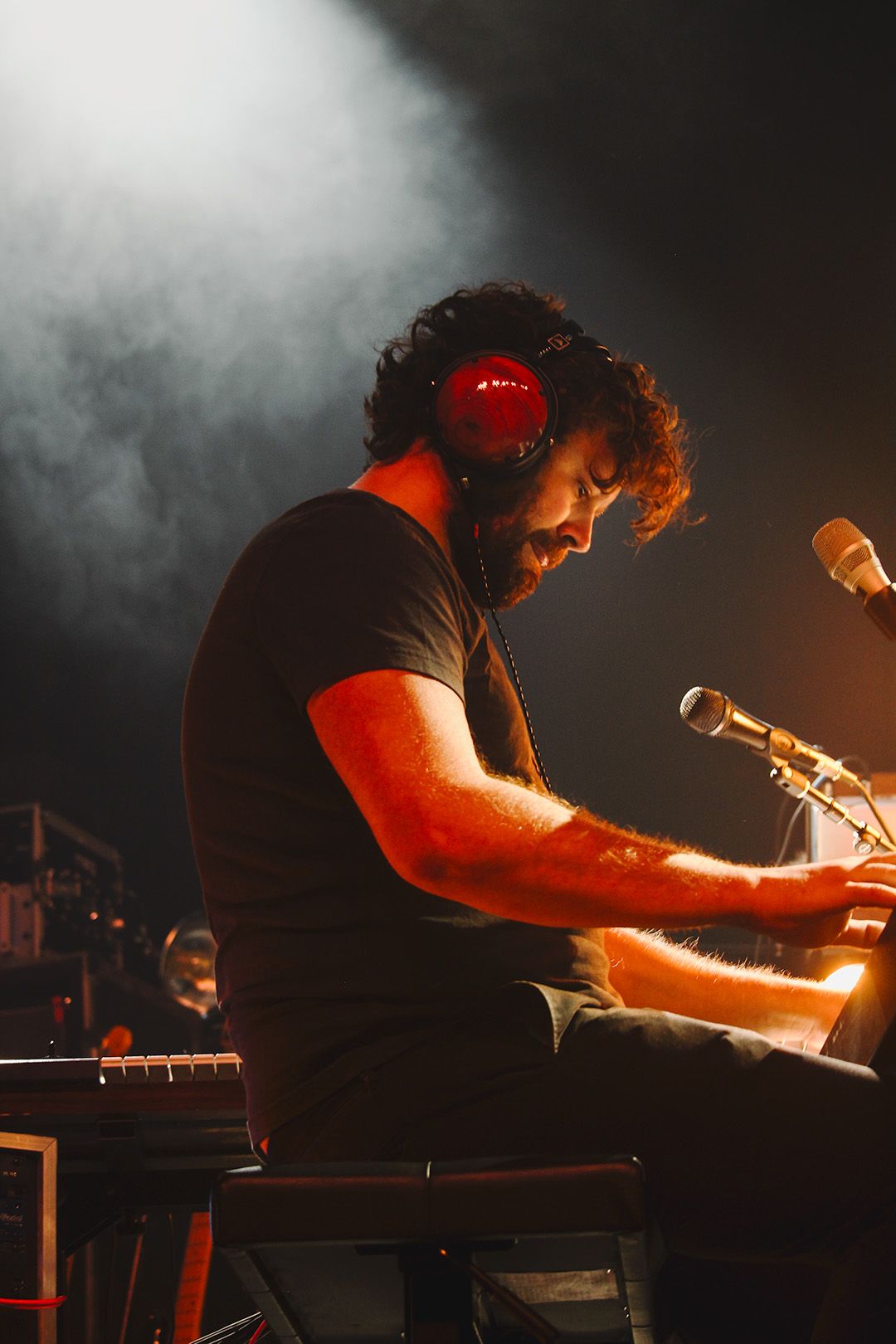 Bearded male pianist wearing red headphones performs on stage surrounded by atmospheric smoke and warm lighting.