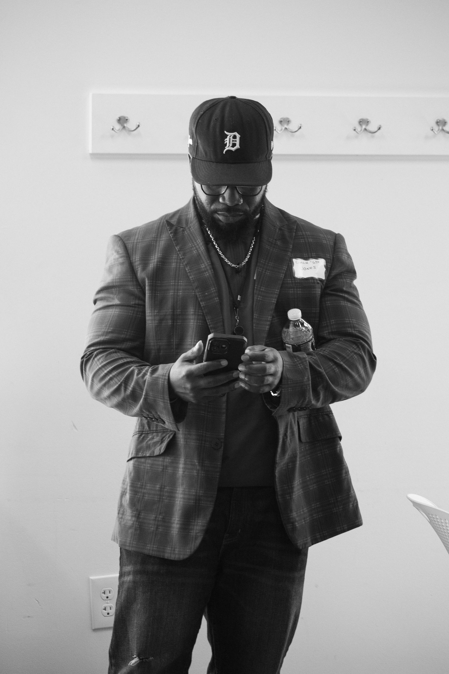 Black and white photo of a man in a plaid blazer and Detroit Tigers cap looking at his phone.