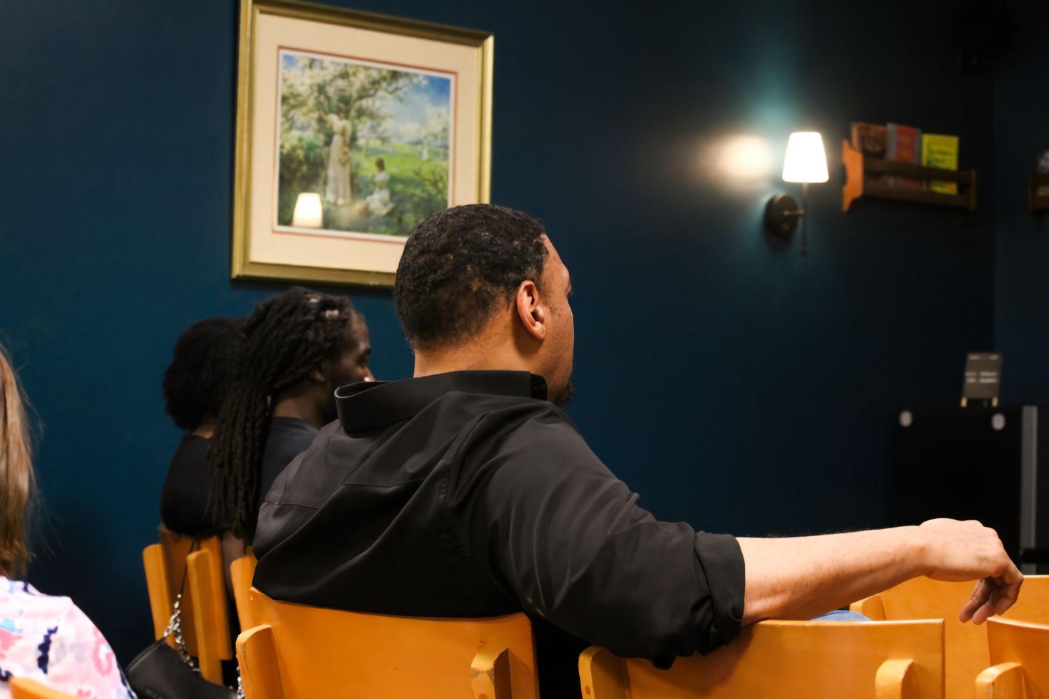People seated in wooden chairs facing a painting on a dark blue wall, lit by a wall sconce in an indoor setting.
