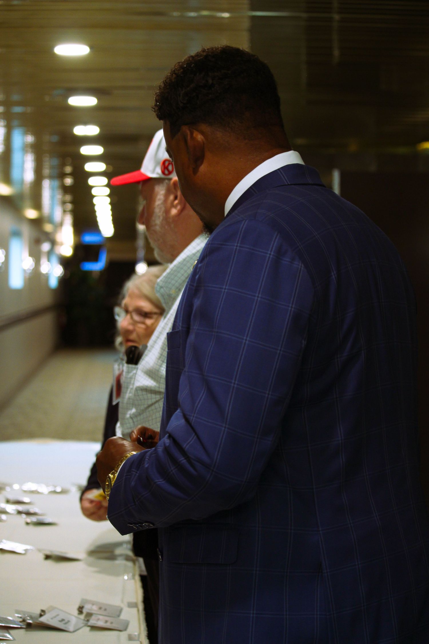 Black man in blue plaid suit standing at a registration table with an elderly woman nearby.