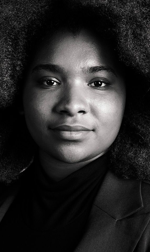 Black and white portrait of a young woman with natural afro hair, wearing a blazer, looking directly at camera.