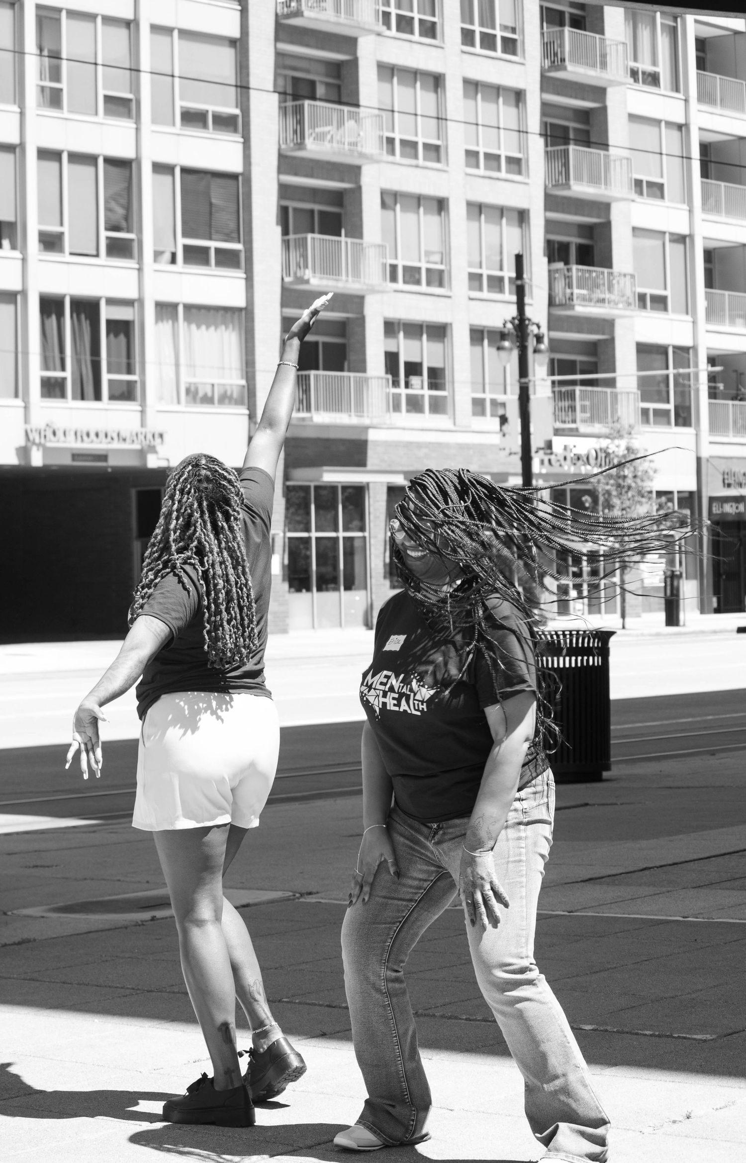 Two people with braided hair dancing outdoors in black and white photo near urban apartment building.