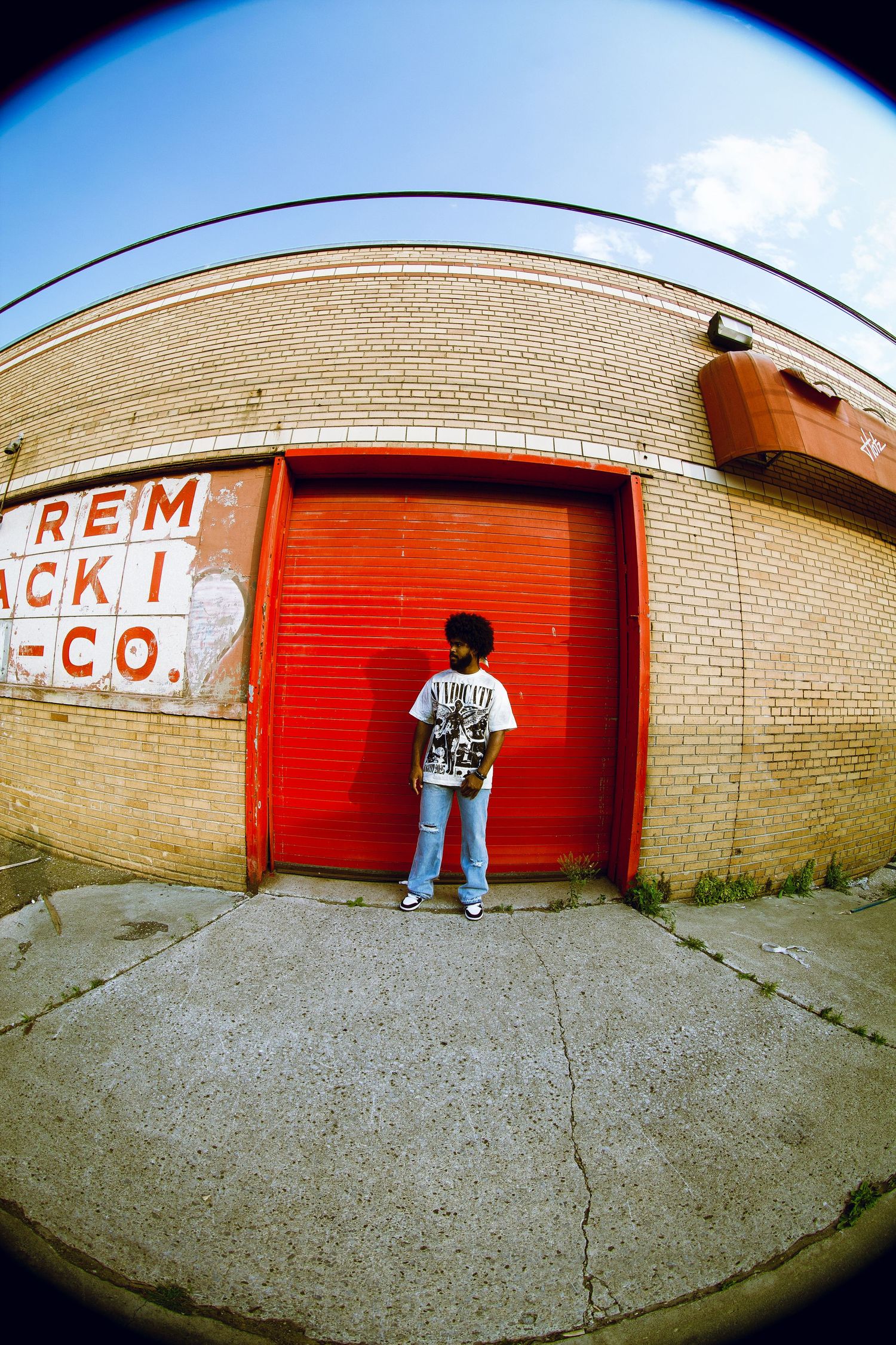 Person in white tee and jeans stands before red garage door on urban street, fisheye perspective.