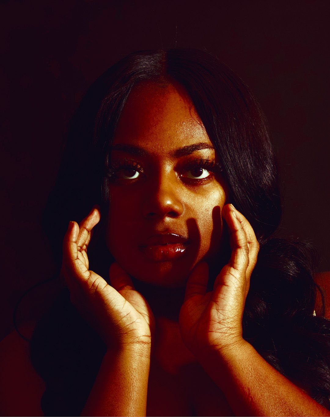 A young Black woman poses with hands framing her face against a dark background in dramatic portrait lighting.