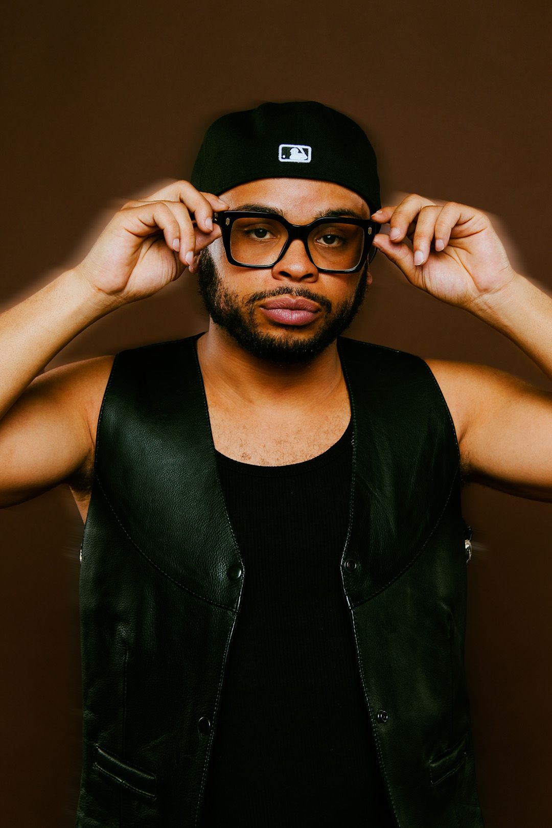 A young Black man in a black leather vest and cap adjusts his glasses against a dark brown background.