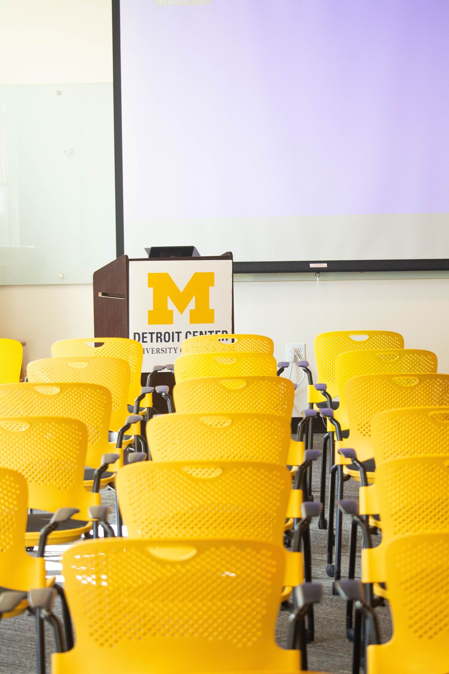 Empty University of Michigan Detroit Center classroom with bright yellow chairs and a podium displaying the iconic block M logo.