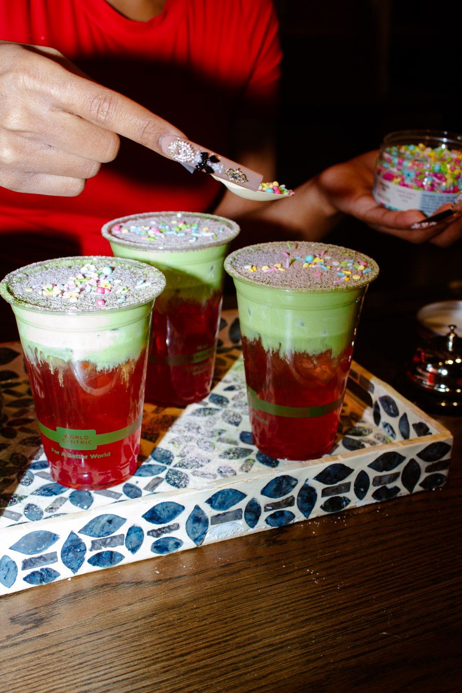 Three layered red and green matcha drinks being decorated with colorful sprinkles on a tray.