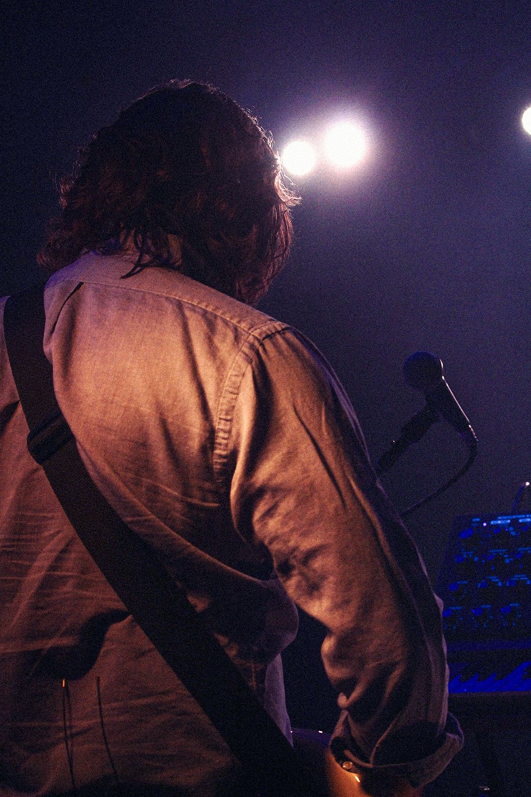 Back view of long-haired guitarist on stage with bright spotlights and microphone stand visible.