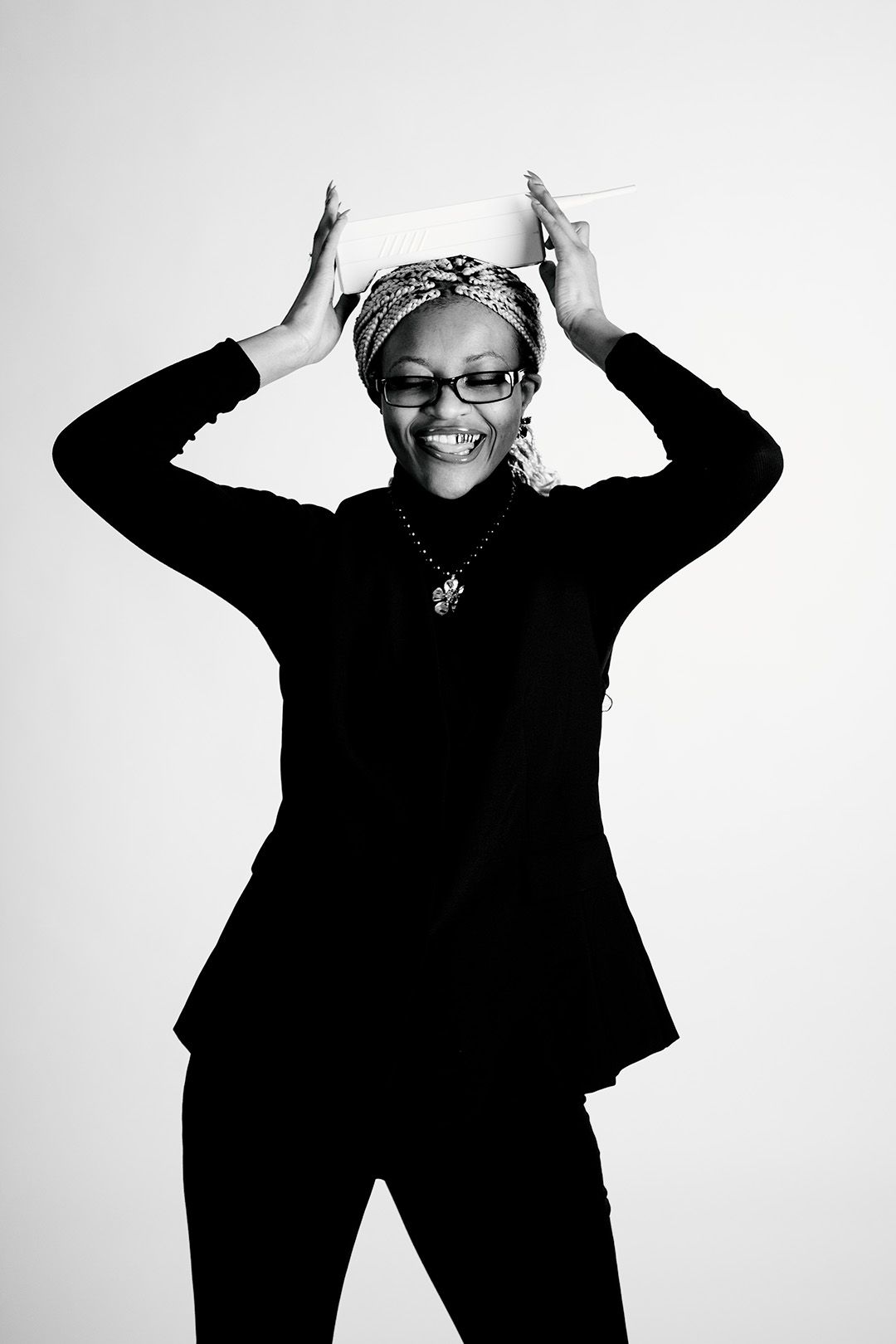 Smiling Black woman in glasses and black suit balancing a white book on her head, black and white photo.