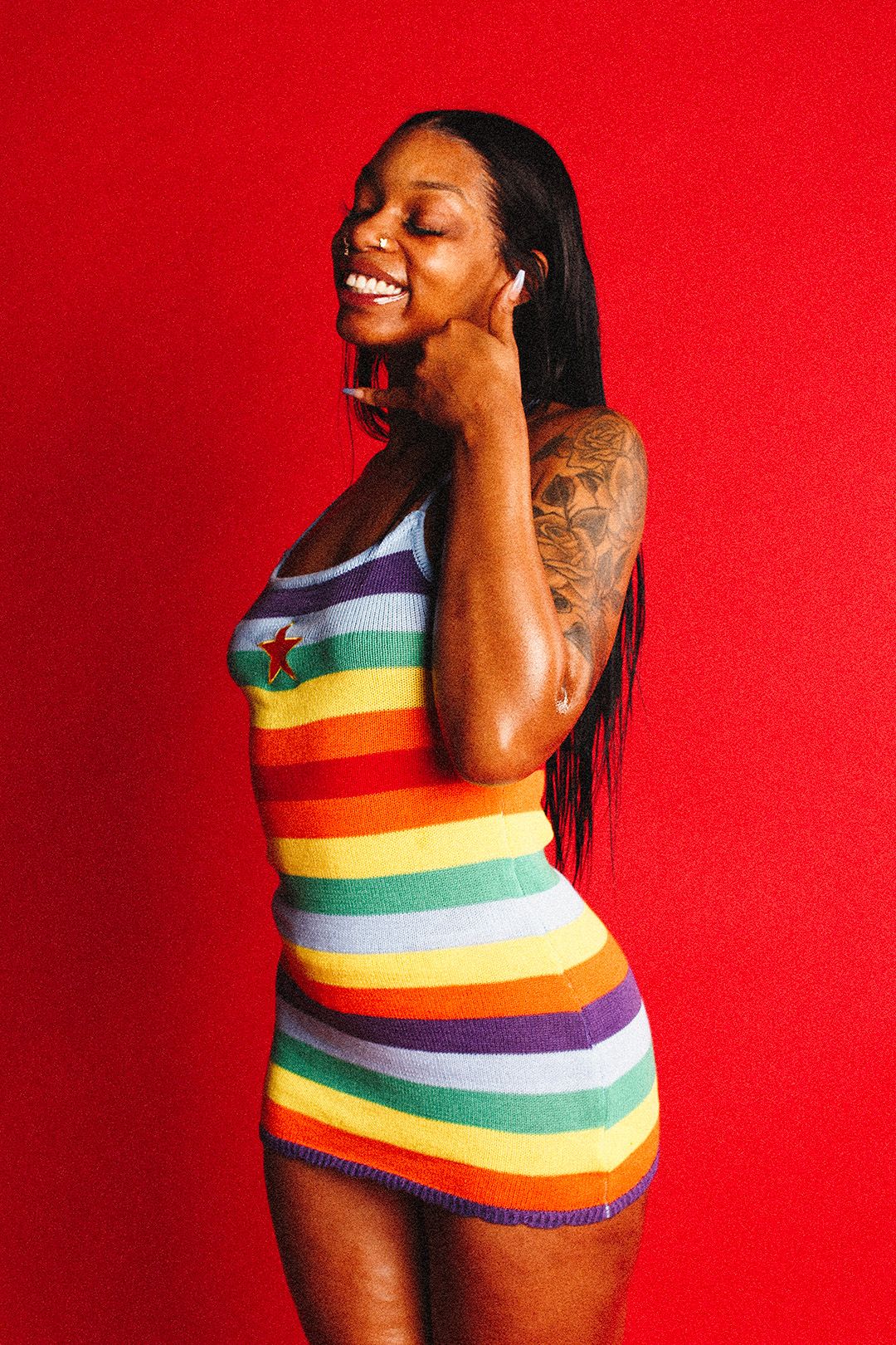 Smiling woman in a rainbow striped dress with tattoos posing sideways on a red background.