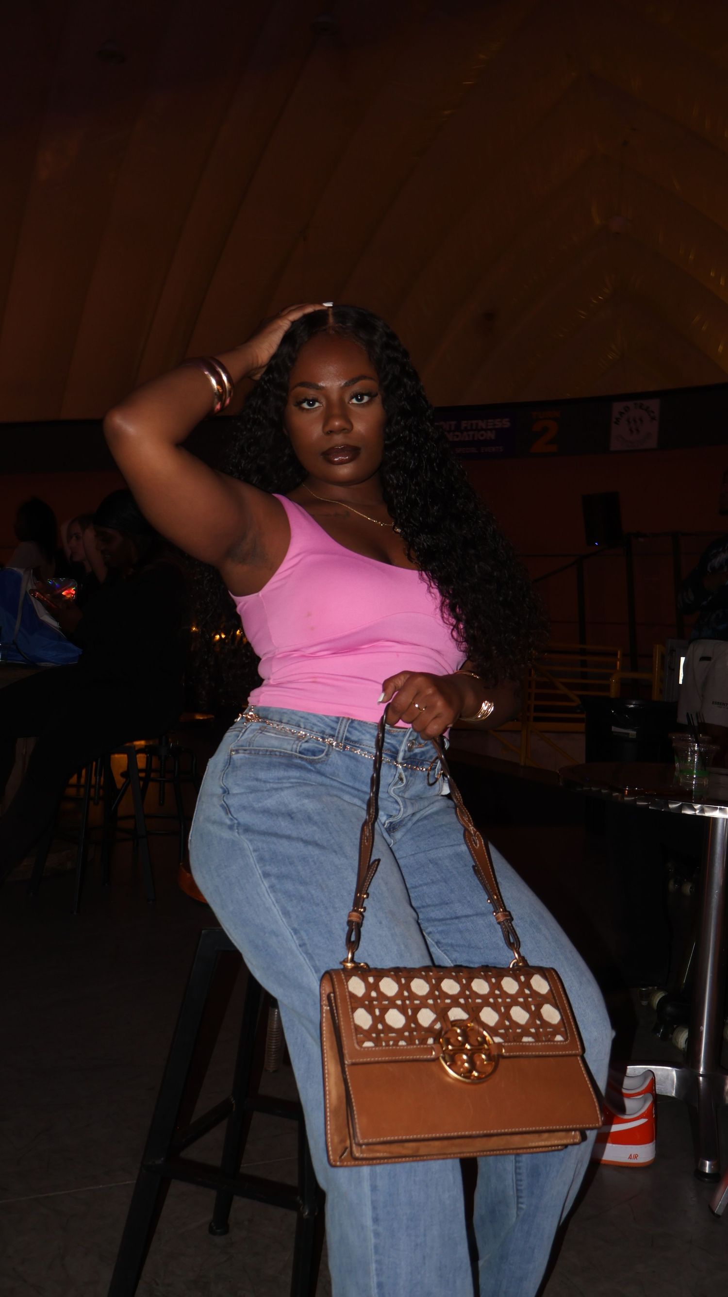 Young Black woman in pink crop top and jeans posing with brown designer handbag at venue.