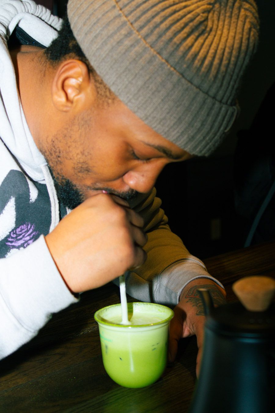 Man in beanie hat sipping an iced matcha latte through a straw at a cafe table.