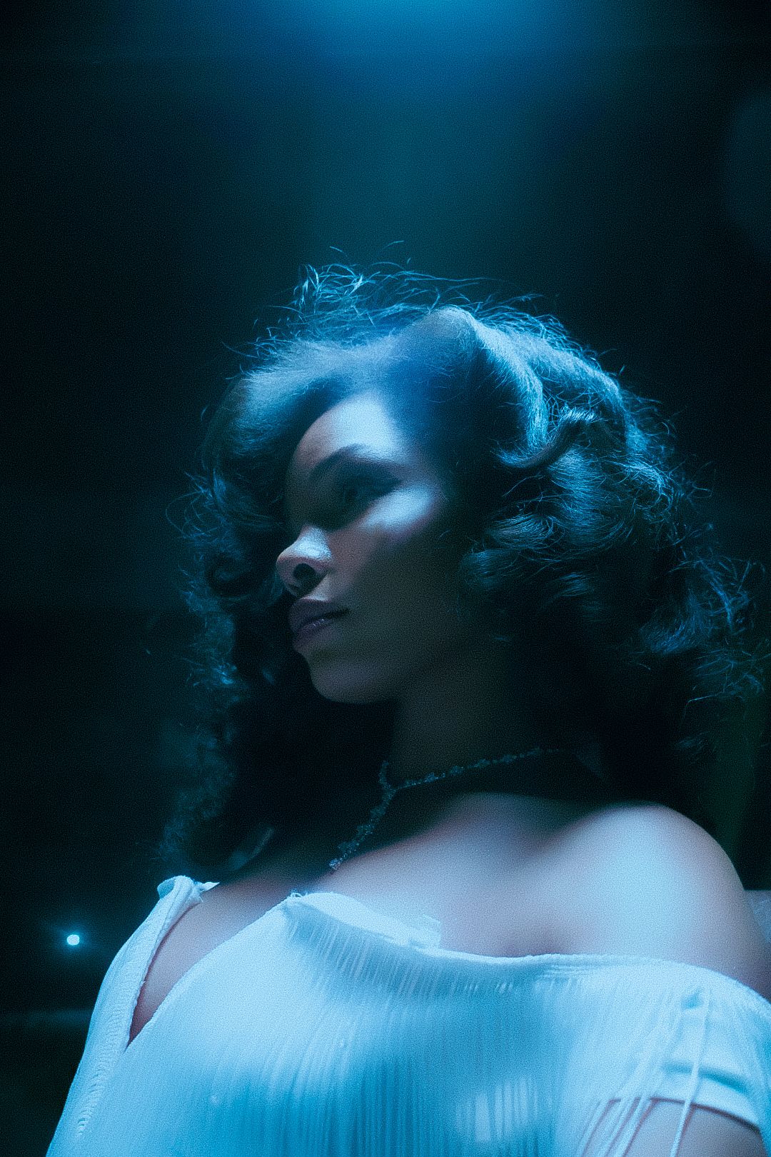 A woman with curly hair poses in a white dress under dramatic blue studio lighting against a dark background.