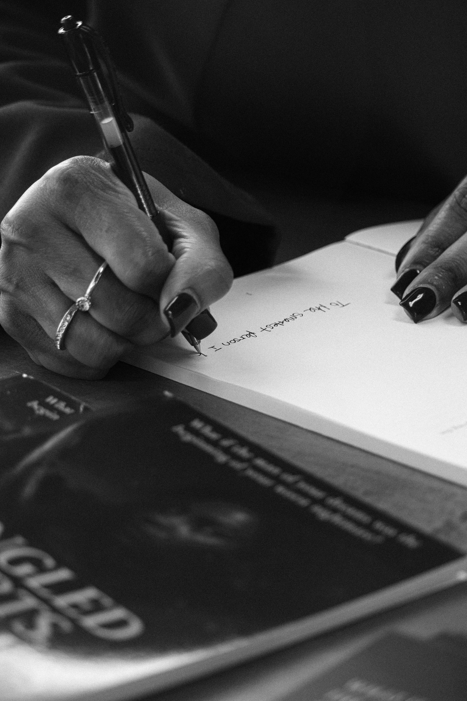 Close-up black and white photo of hand with dark nails signing a book with a pen.