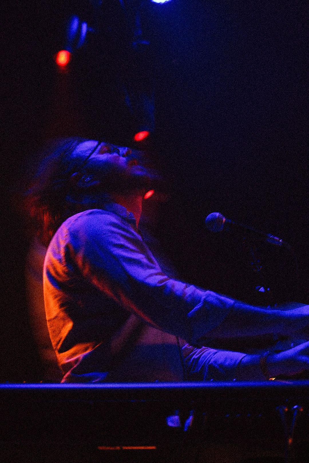 Musician playing keyboard on stage illuminated by blue and red stage lighting during live performance.