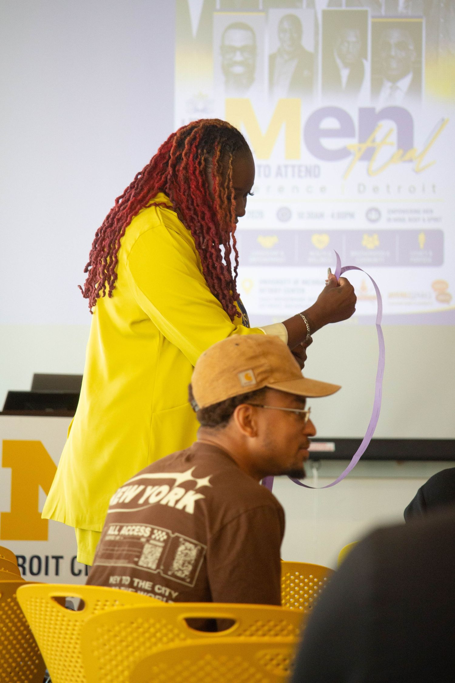 Woman with red braids in yellow outfit placing ribbon on seated man at Mental Health Detroit conference.