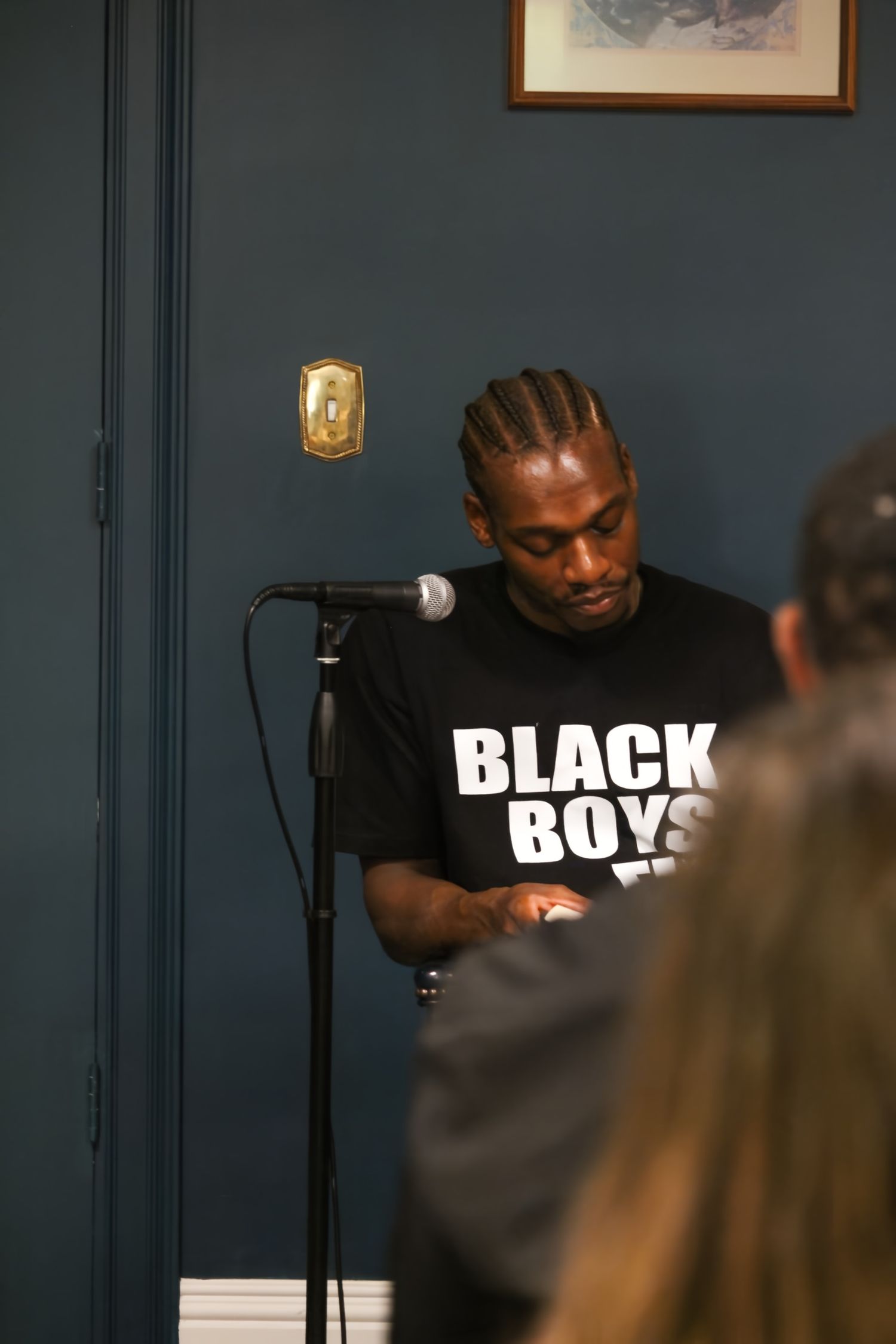 Man in Black Boys t-shirt speaking at a microphone stand indoors before an audience.
