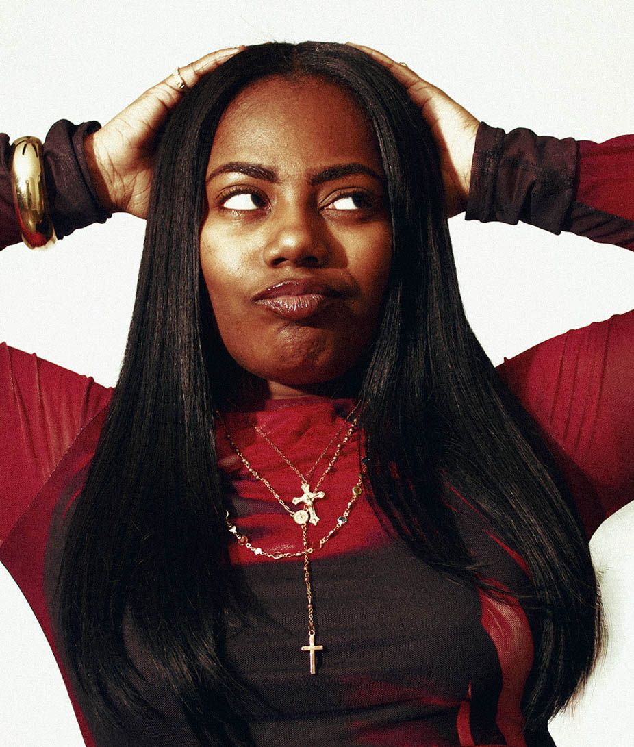 Young woman with long straight black hair wearing a red jacket and gold cross necklace poses with hands behind head.