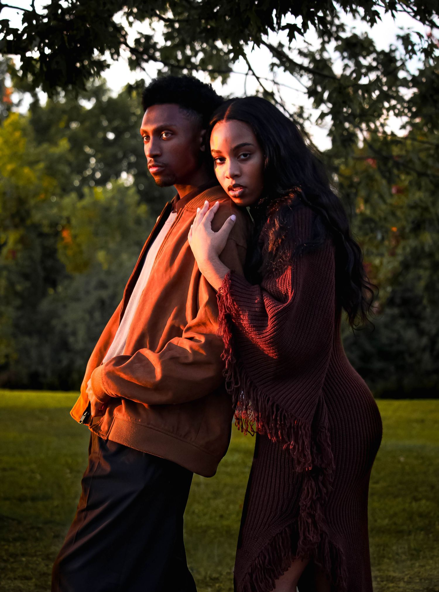 Sunset photo of a fashionable couple in rust and brown outfits posing back to back outdoors.