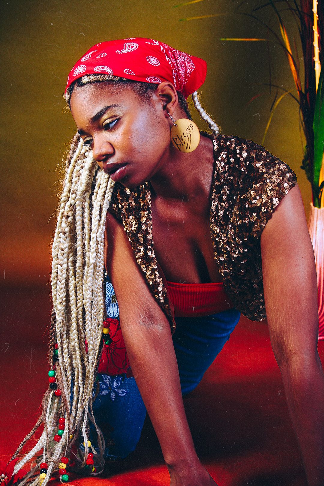 Young Black woman with long blonde locs and red bandana poses confidently in gold sequin top and blue skirt.