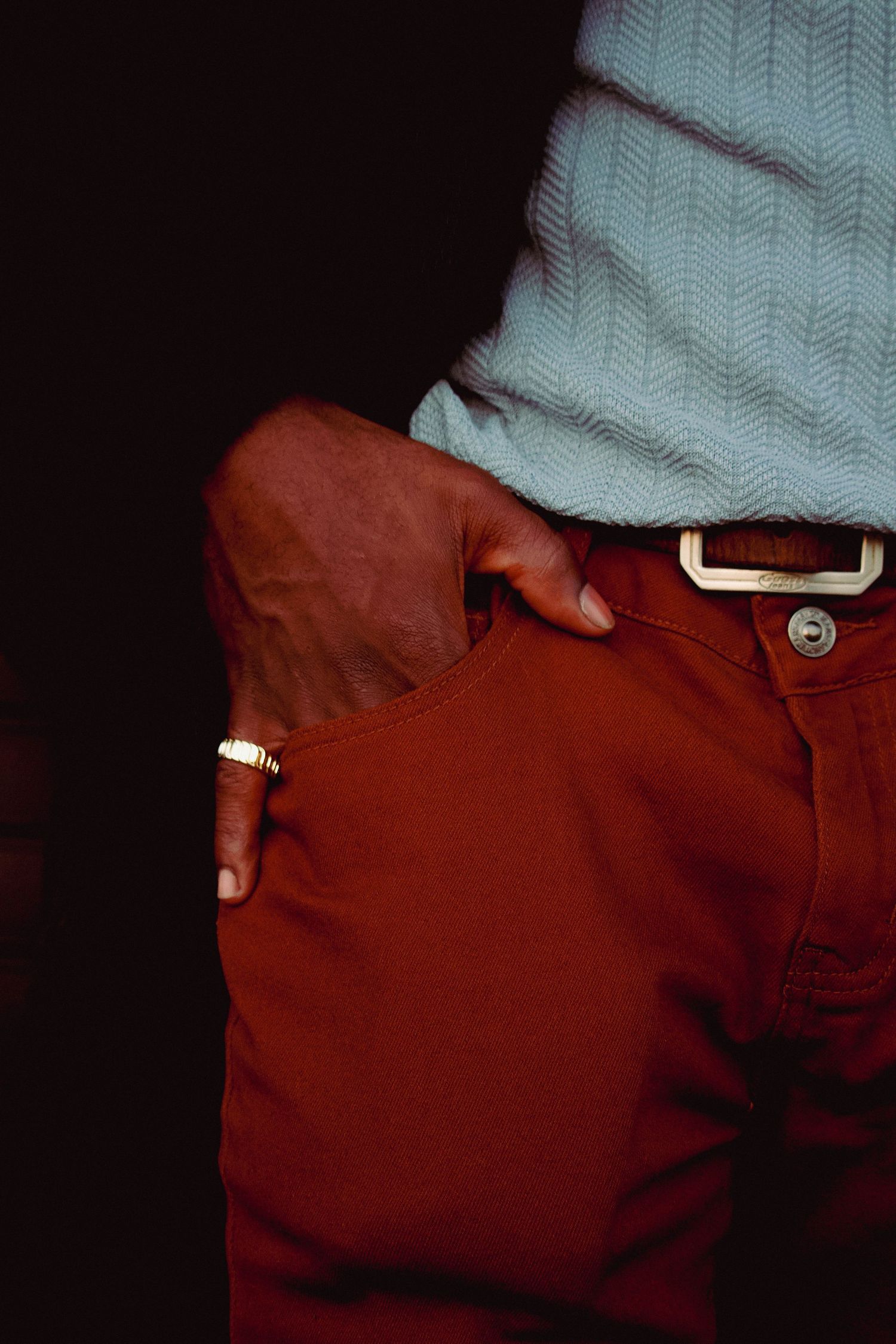 Close-up of a man's hand in red pants pocket, wearing a gold ring, with a light blue knit sweater and brown belt.