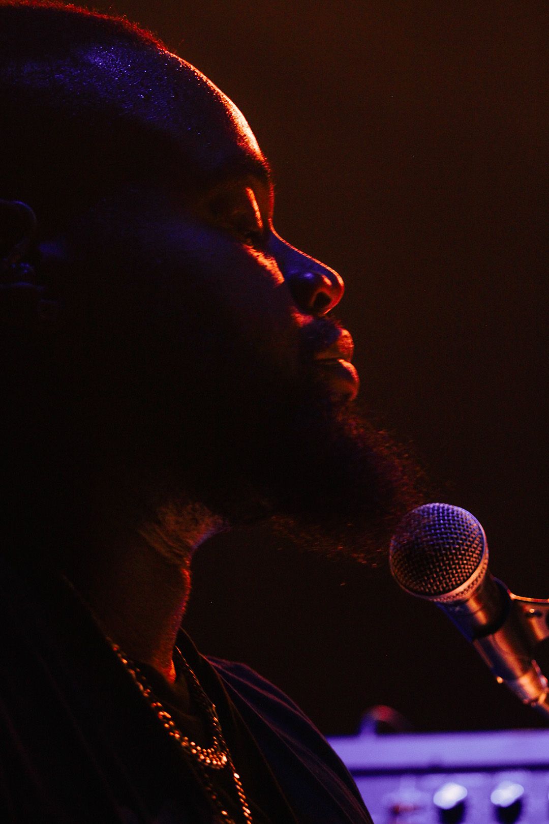 Close-up profile of bearded Black male vocalist performing at microphone under warm red stage lighting.