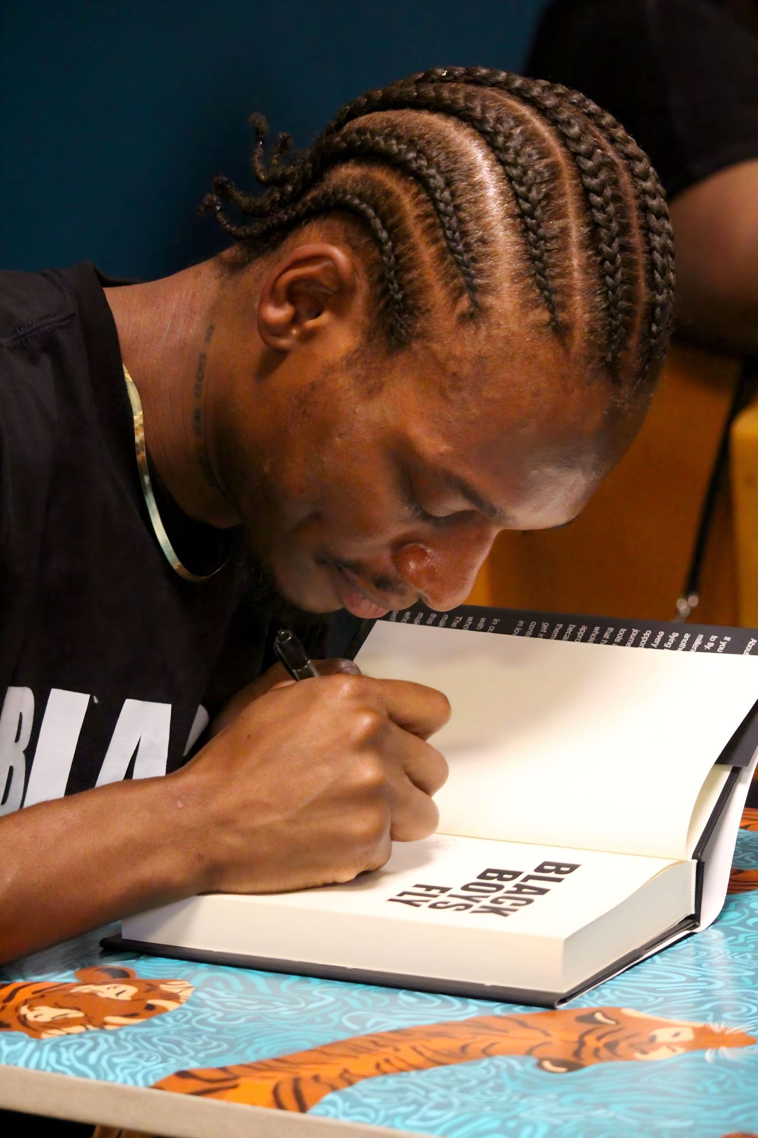 Young man with cornrow braids signing a book at a table with tiger print design.