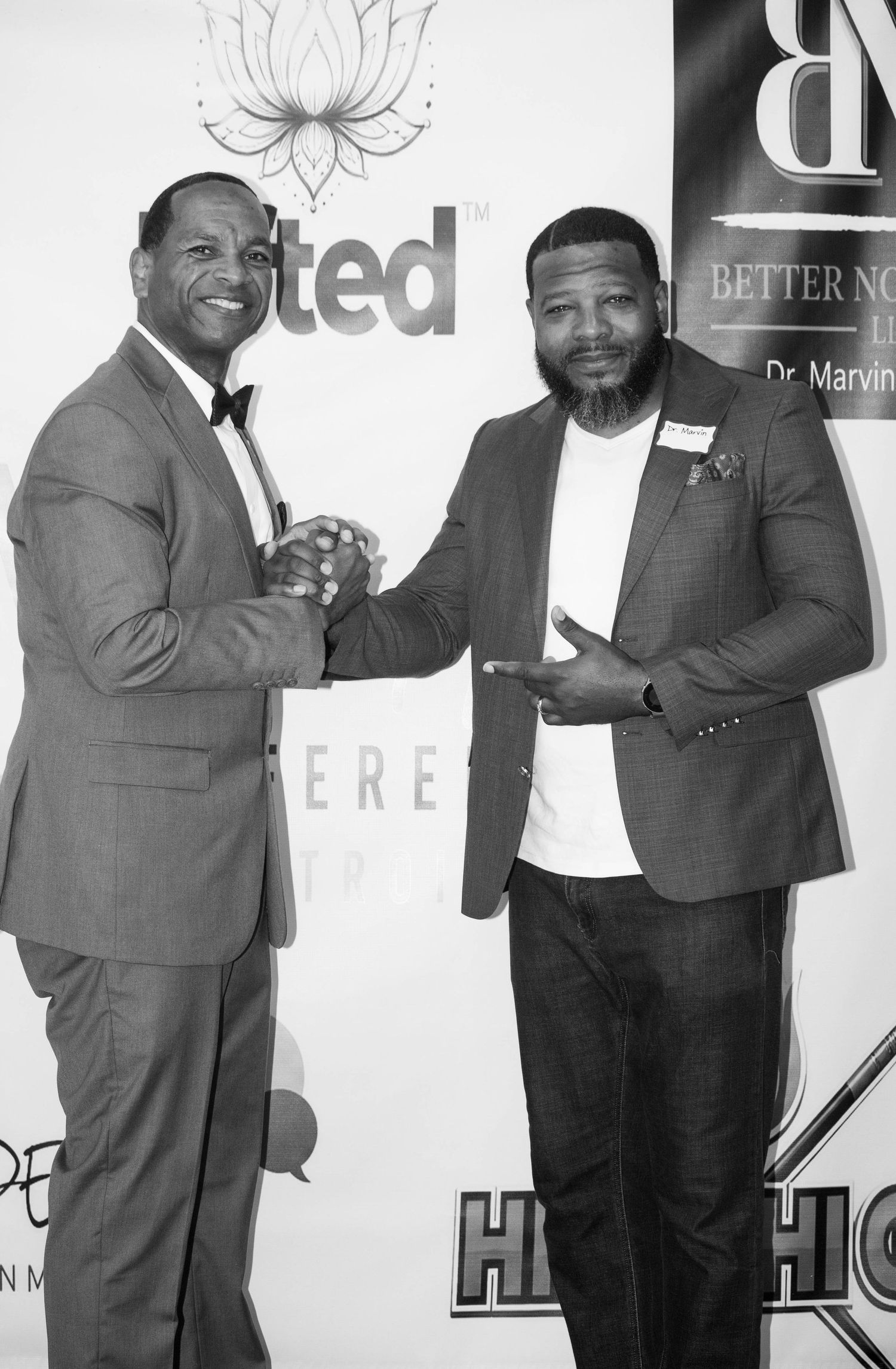 Two men in suits shake hands and pose together on a black and white event backdrop.