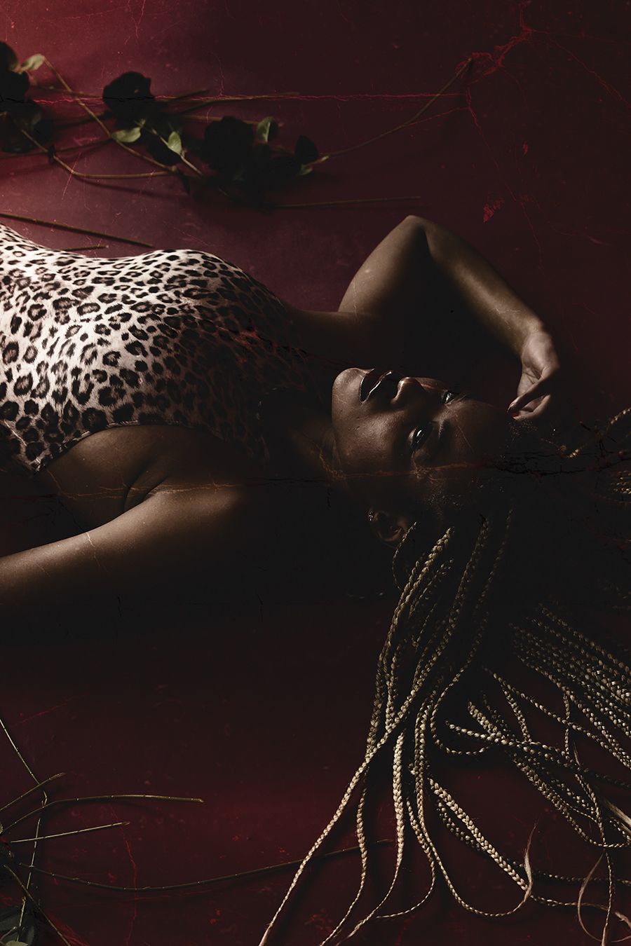 Black woman with long braids in leopard print bodysuit lies on red floor surrounded by dark roses.
