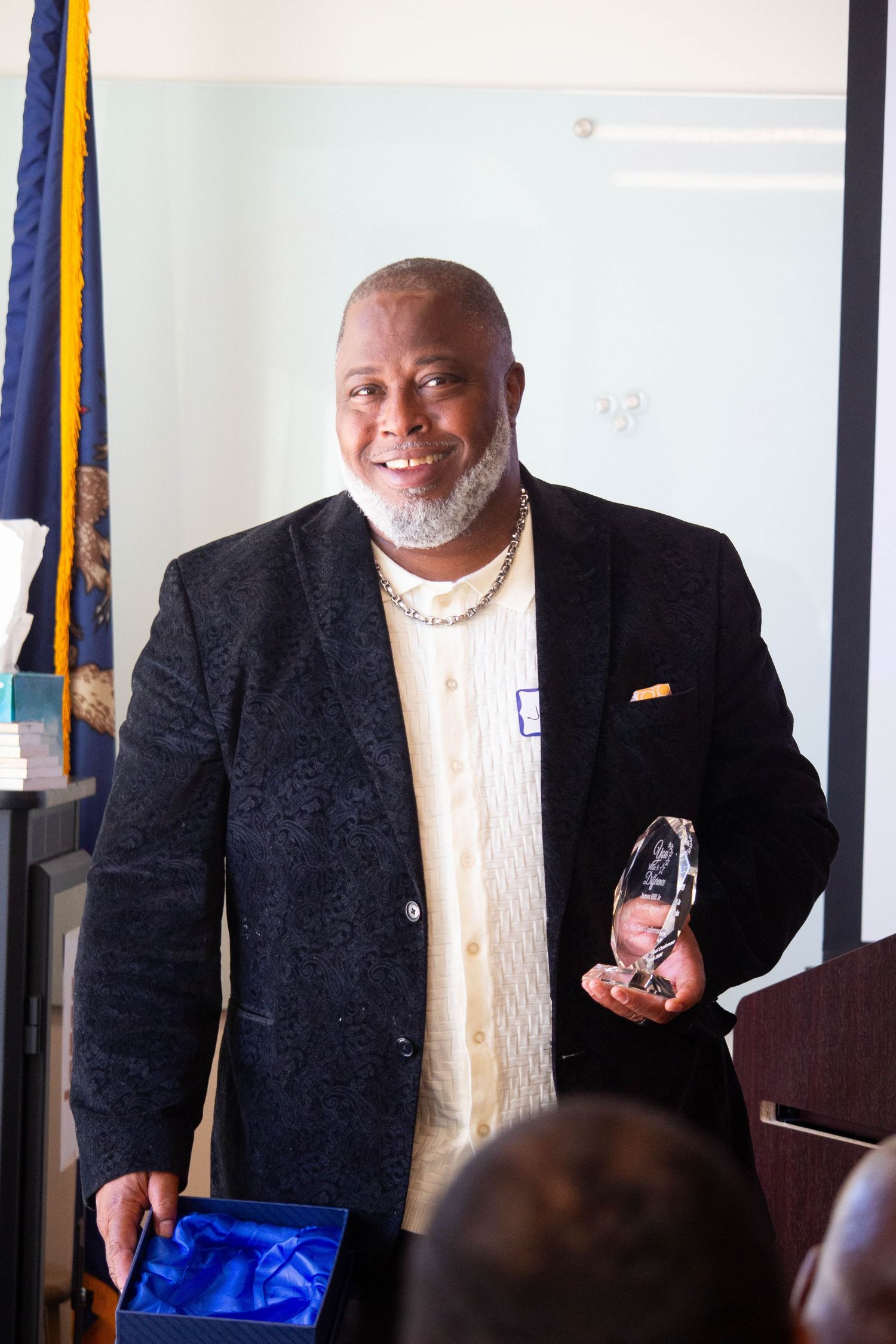 Smiling man in black blazer holding a crystal award trophy at an indoor recognition ceremony.
