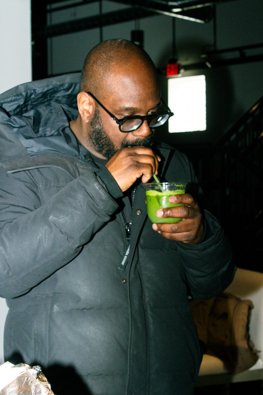 Bald man with glasses and beard drinking a green matcha smoothie through a straw.