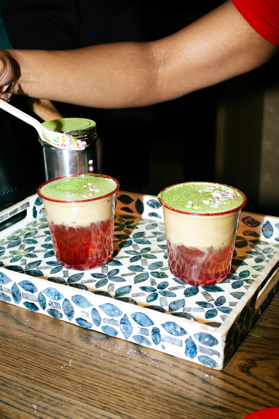 Two layered matcha drinks being garnished with green powder on a decorative tray.