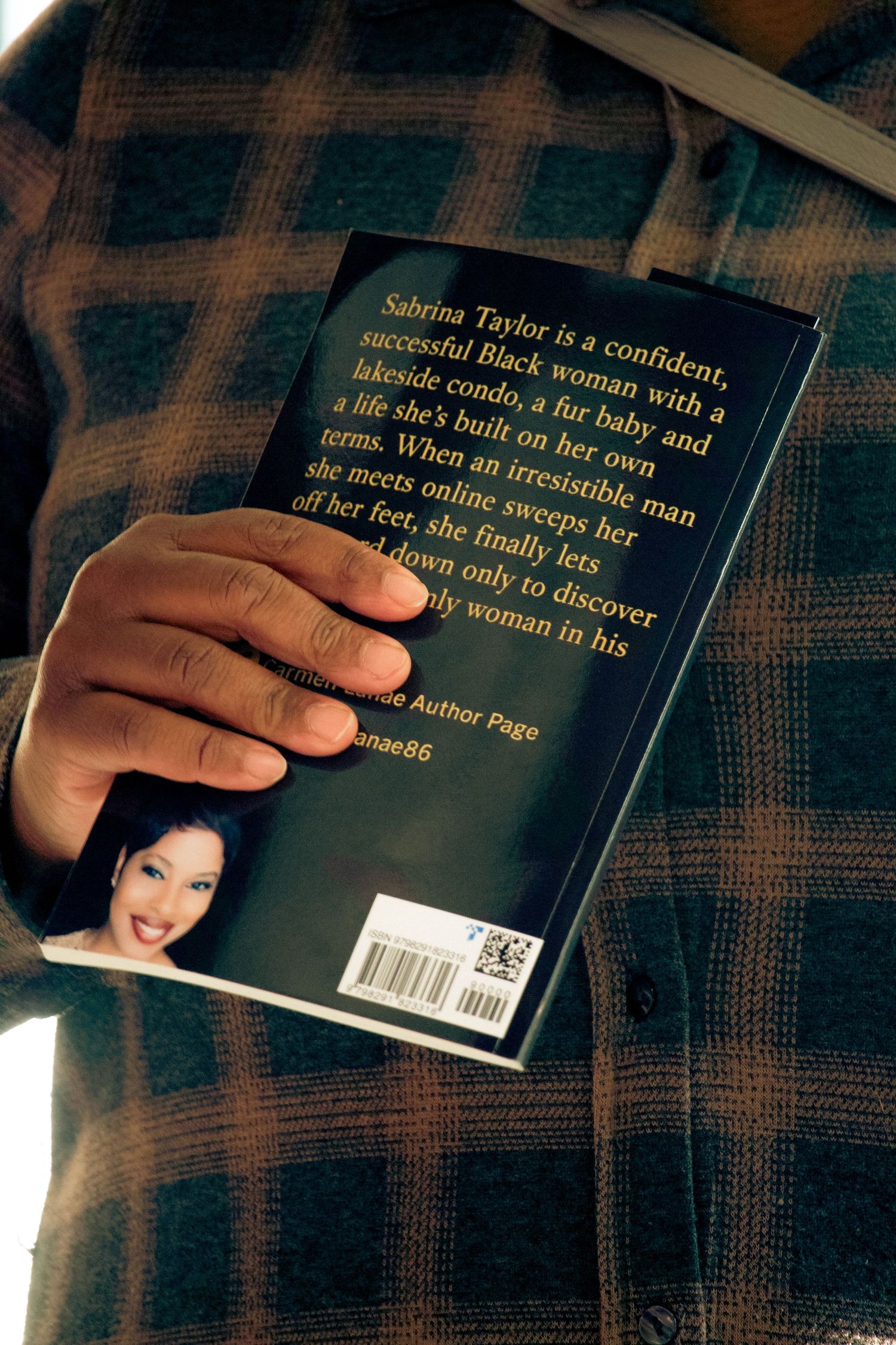 Person in plaid shirt holding a novel with back cover text about Sabrina Taylor, a confident Black woman.