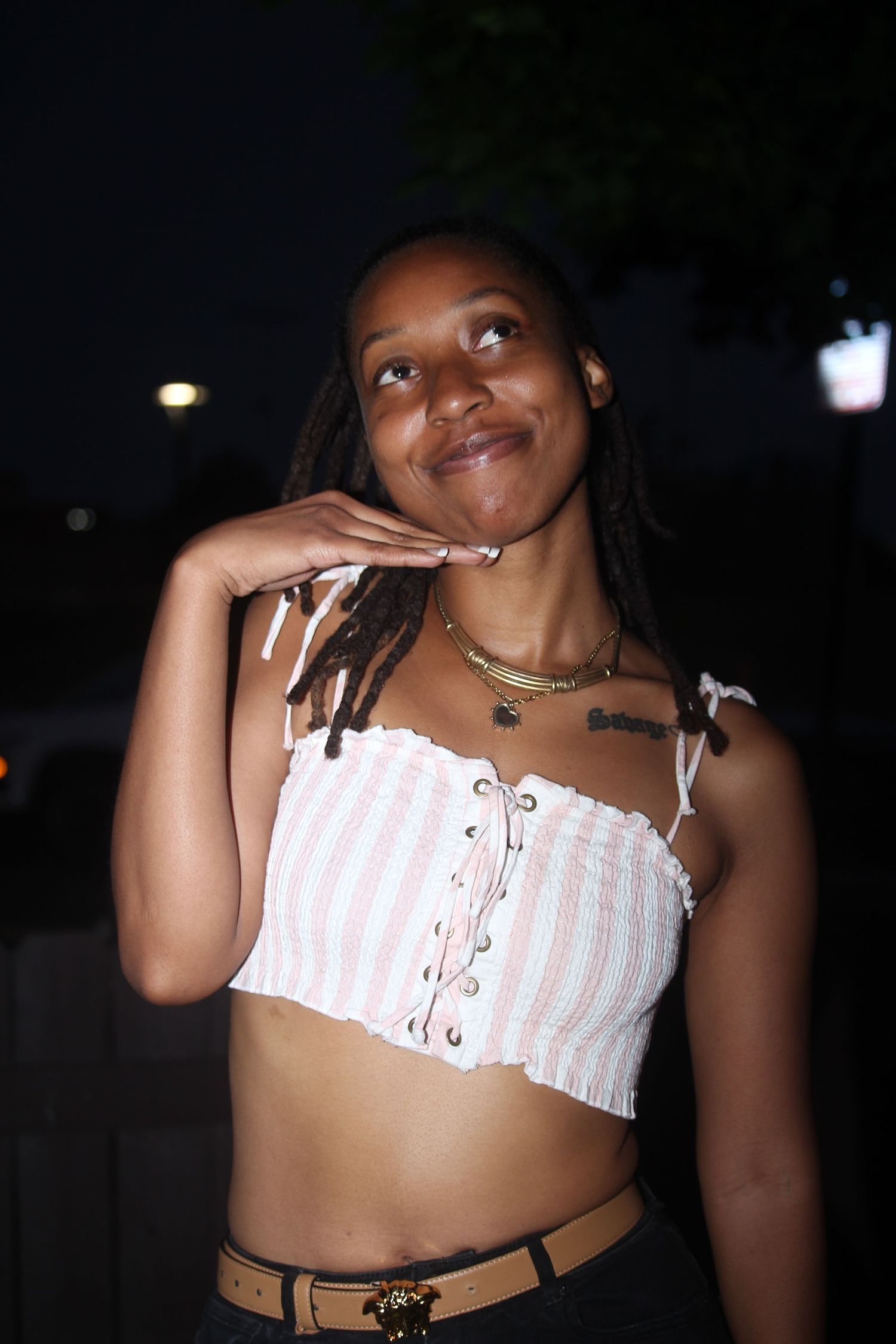 Young woman in striped crop top poses thoughtfully at night, wearing layered gold necklaces.
