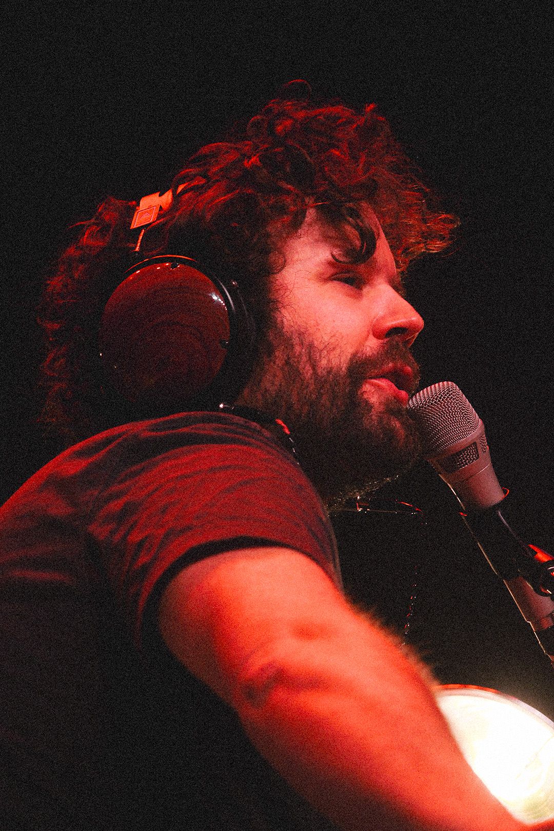Curly-haired male musician wearing headphones singing into microphone under warm red stage light.