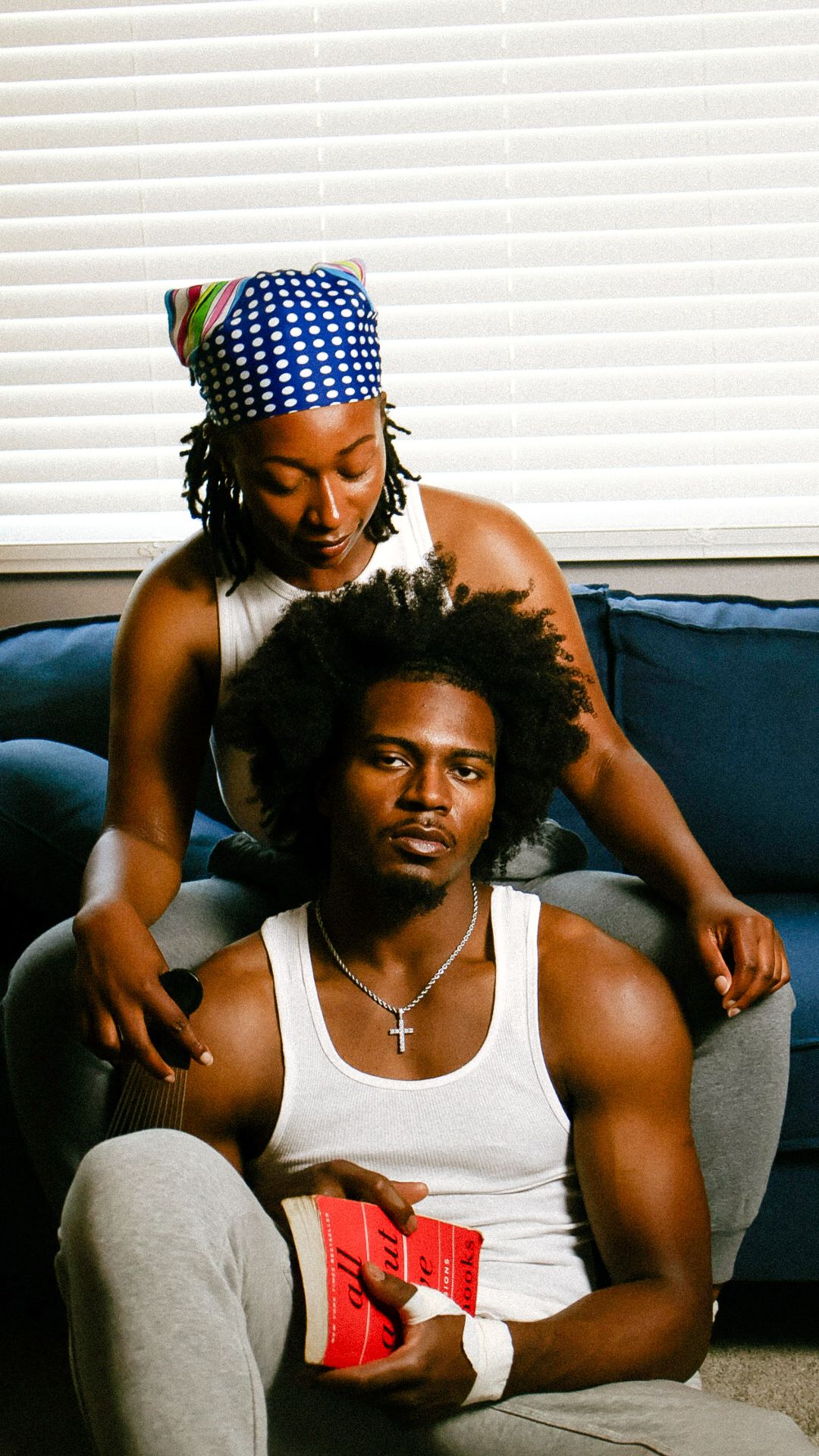 A woman braids a mans hair as he sits on the floor holding a red book near a blue couch.