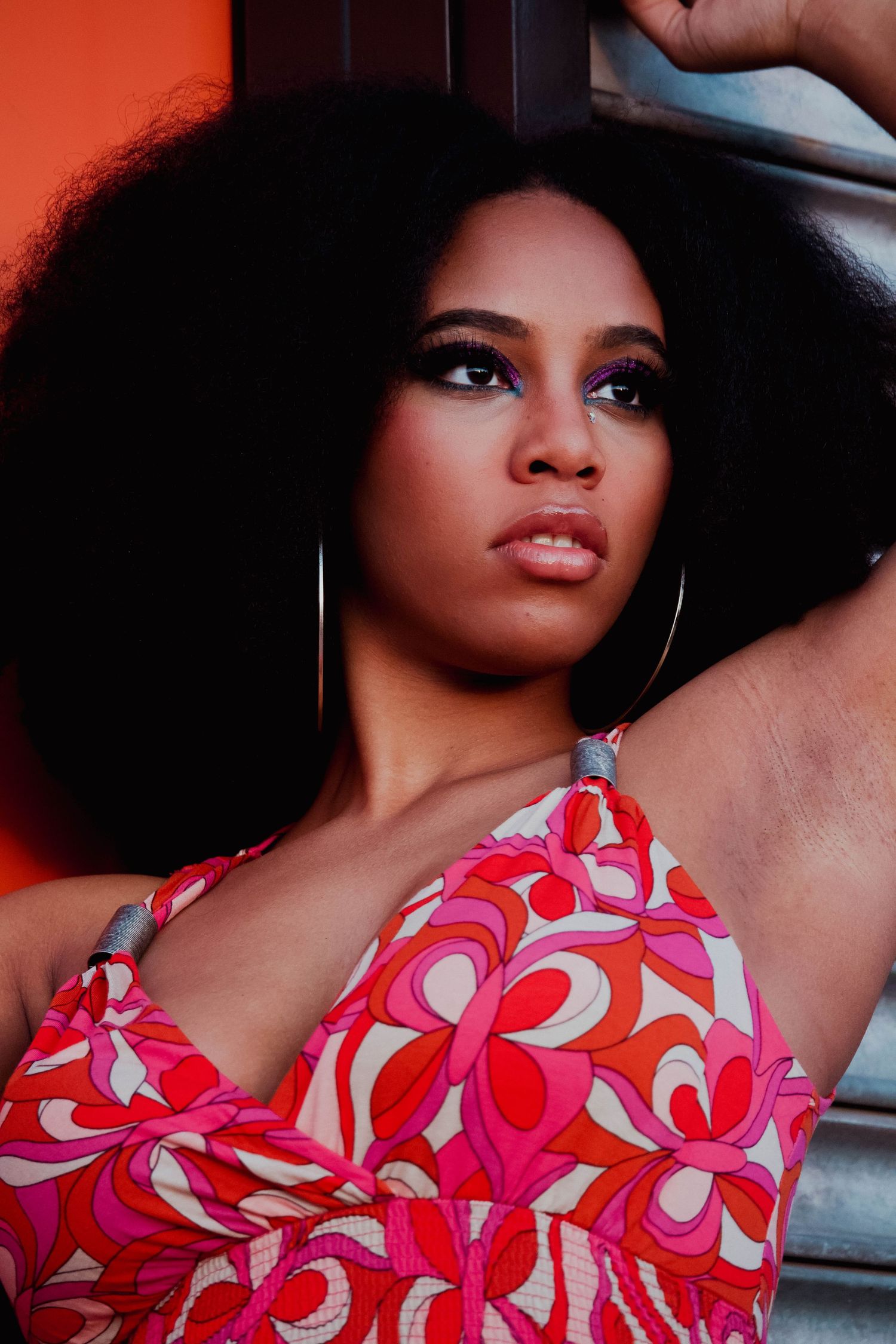 A woman with a large afro and hoop earrings poses in a colorful pink floral dress against an orange wall.