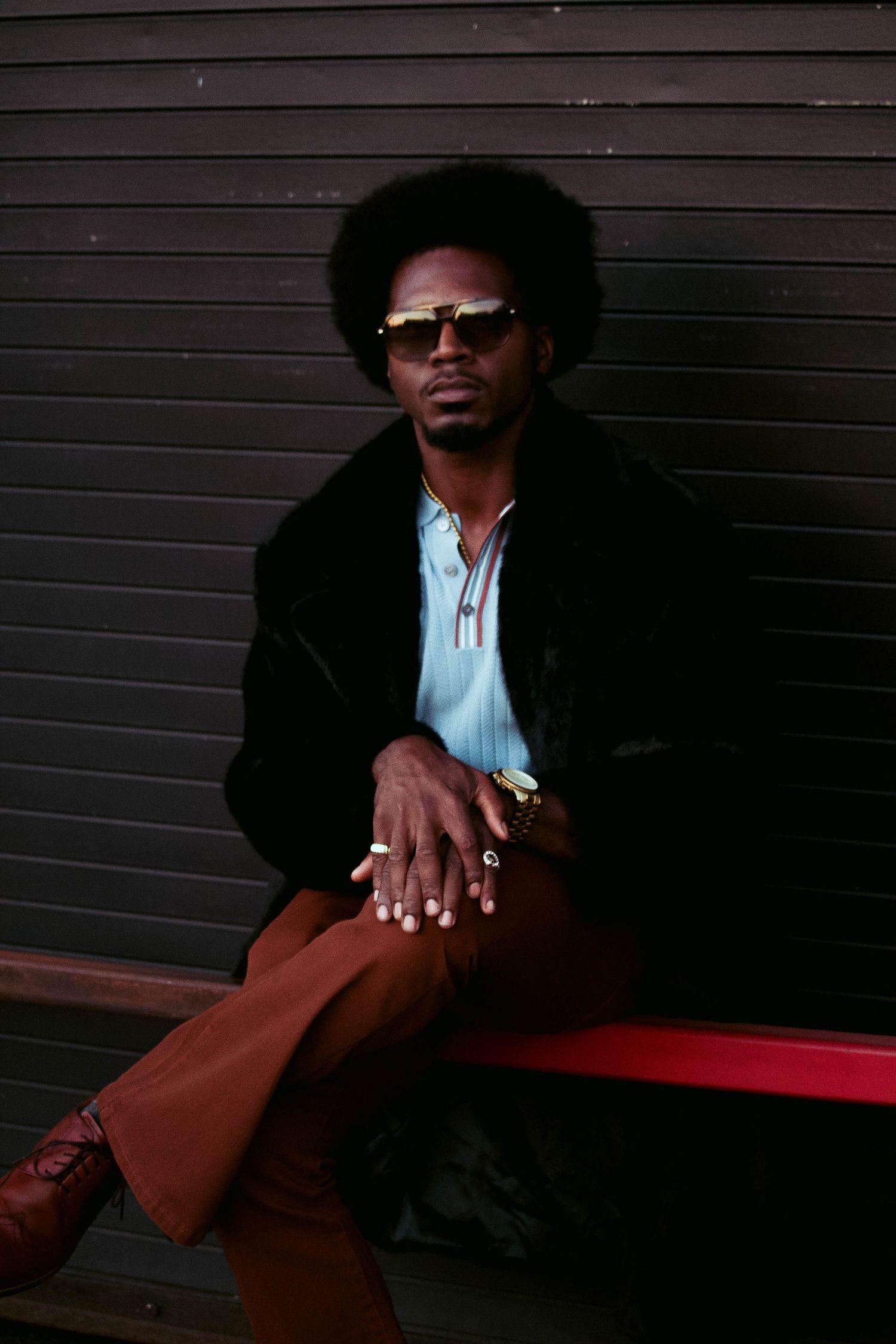 Stylish man with afro wearing sunglasses, black fur coat, brown flares, sitting on red bench against dark wall.