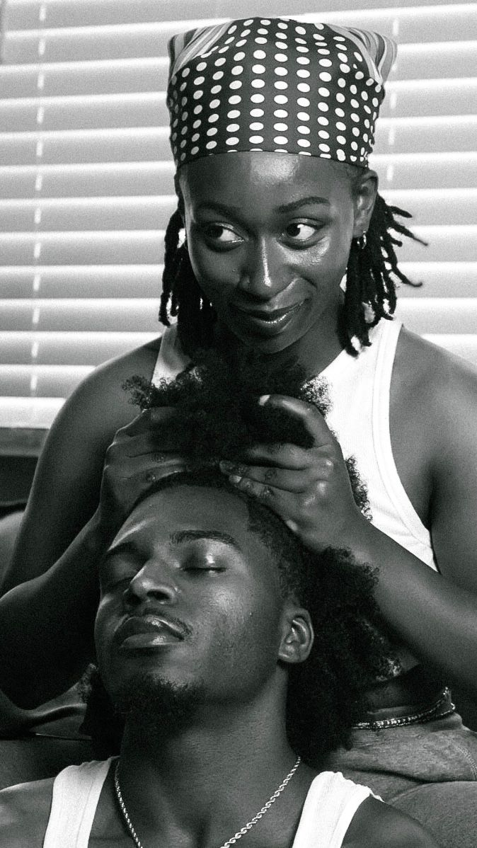 Black and white photo of a woman in a polka dot headscarf styling a man's natural hair indoors.