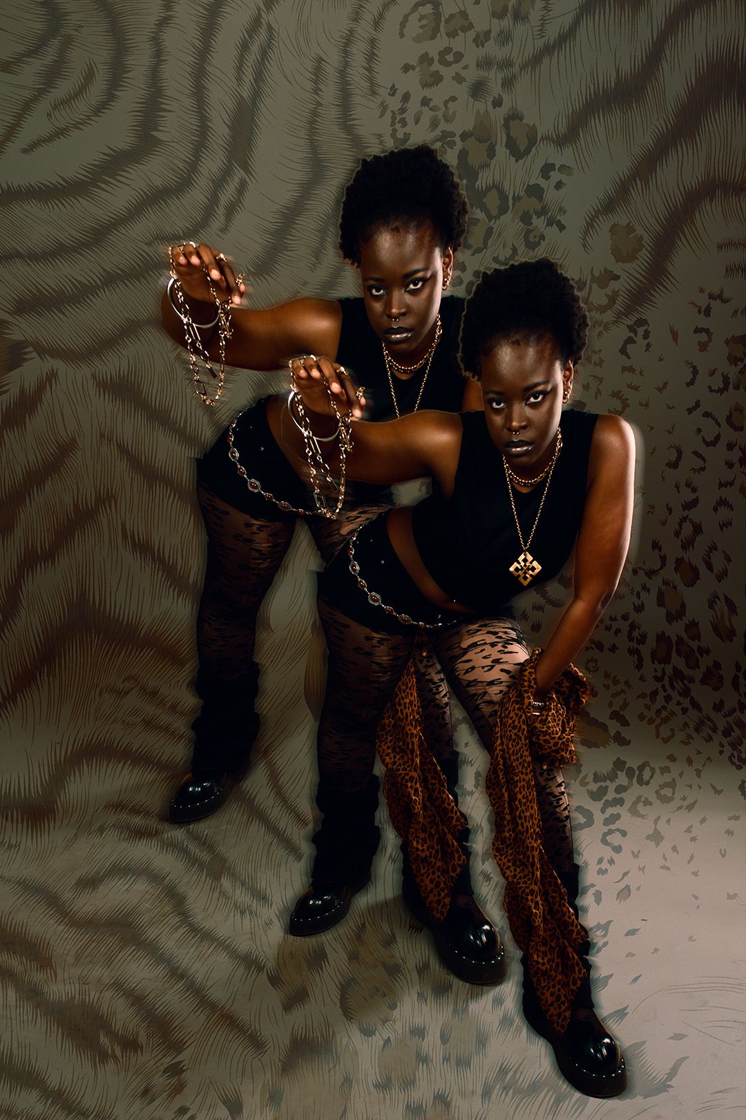 Two dark-skinned women pose dramatically in leopard print outfits holding chains against an animal print backdrop.
