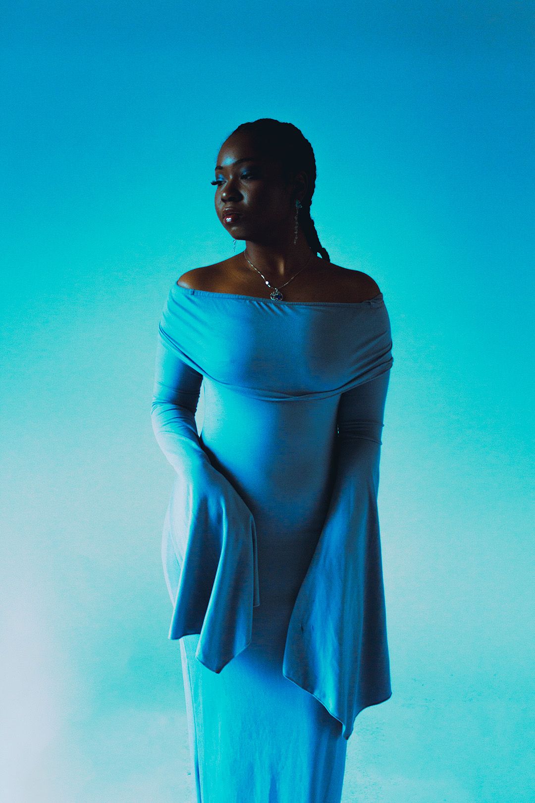 Woman in a blue off-shoulder dress posed elegantly under teal studio lighting.