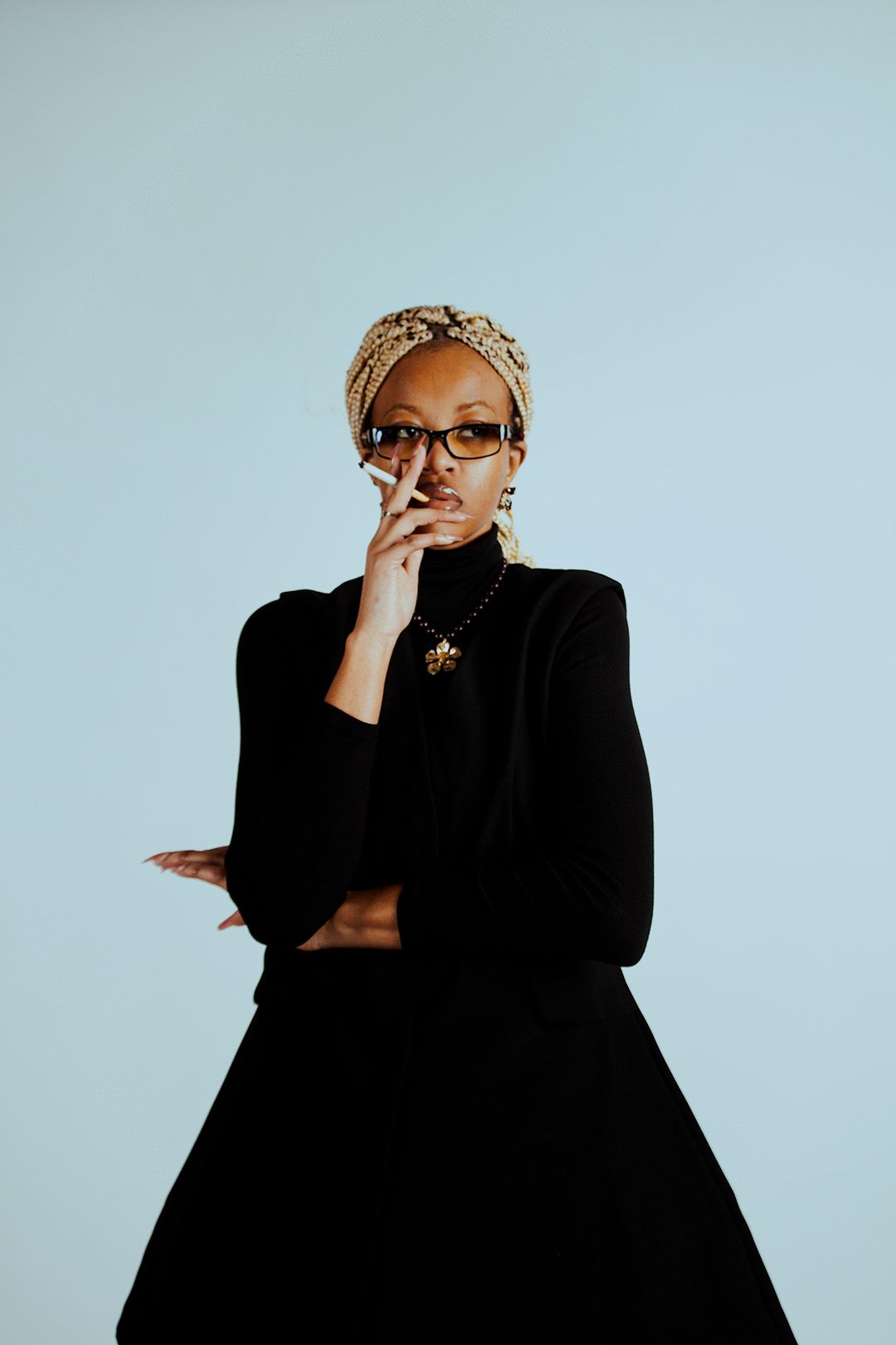 A stylish Black woman in a black outfit and gold head wrap poses thoughtfully against a light blue background.