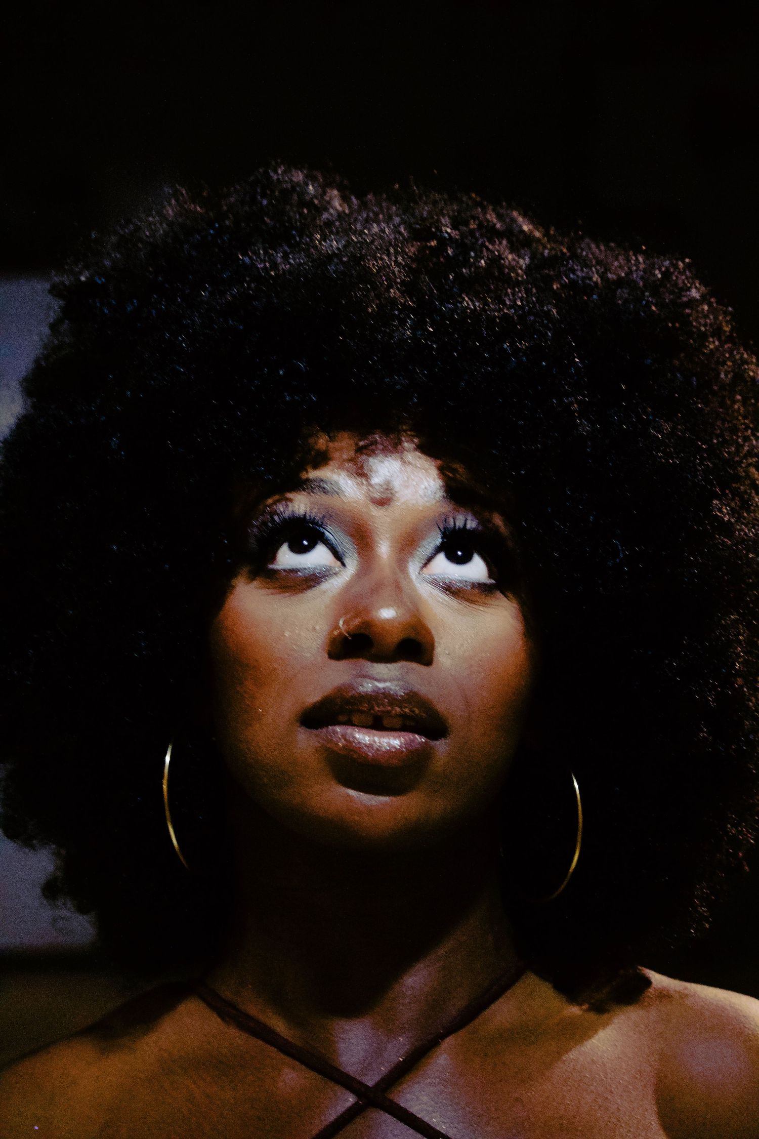 Close-up portrait of a woman with an afro hairstyle, bold eye makeup, and gold hoop earrings against a dark background.
