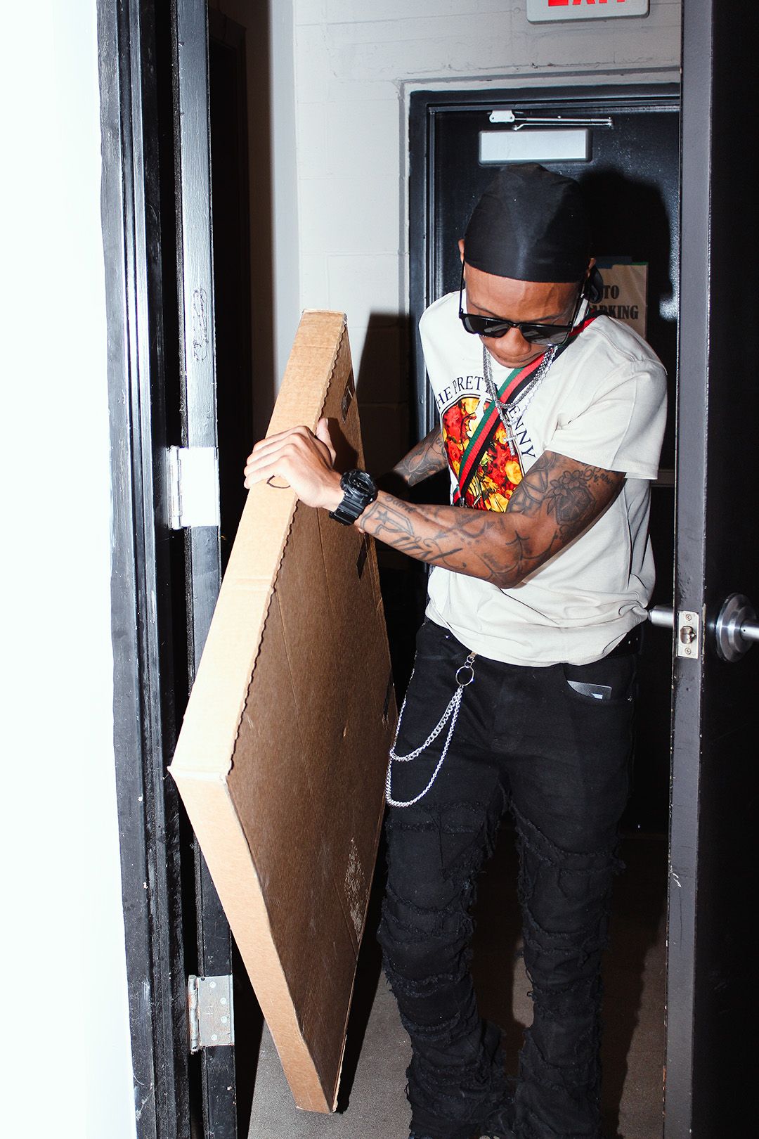 Tattooed man in white graphic tee, sunglasses, and black jeans carrying a large cardboard piece through a doorway.