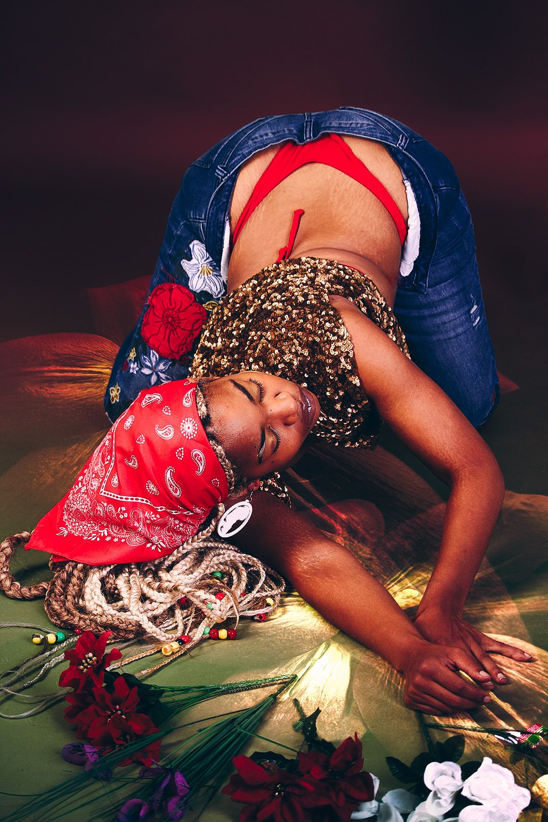 Woman in red top and jeans bowing forward on gold surface surrounded by colorful flowers and beaded braids.