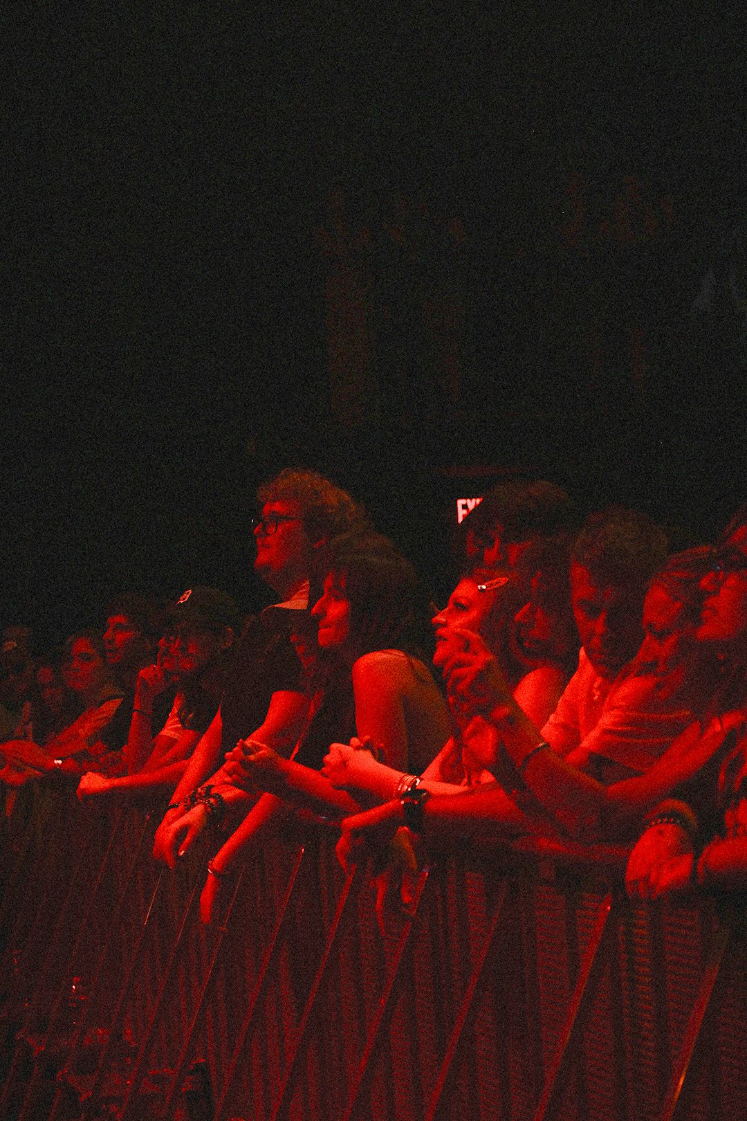 Crowd of concert fans pressed against a barrier, bathed in dramatic red stage lighting at a live music event.